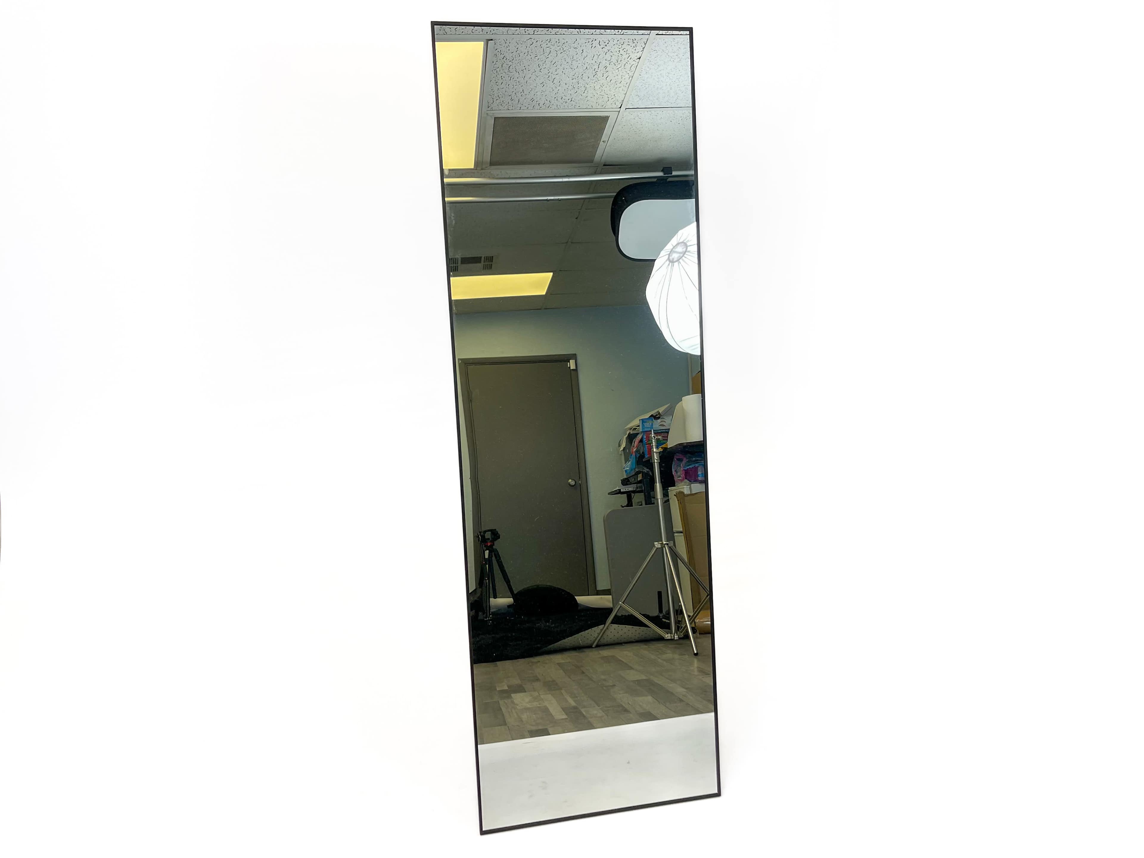 A tall rectangular mirror is mounted on a wall, reflecting a brightly lit room with photography equipment in the background.