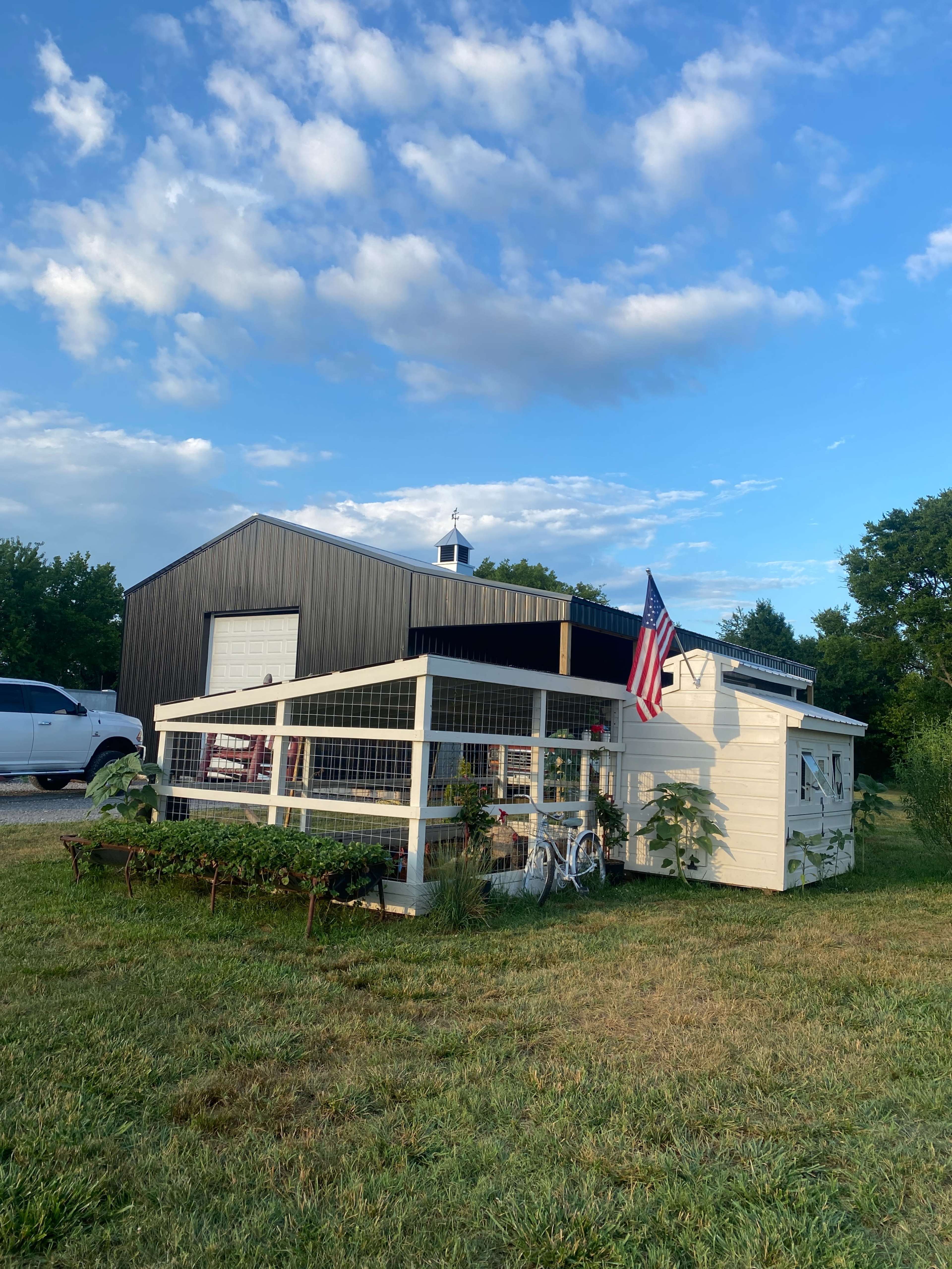 A white shed with a fenced garden area and an American flag stands next to a large black barn under a cloudy blue sky.