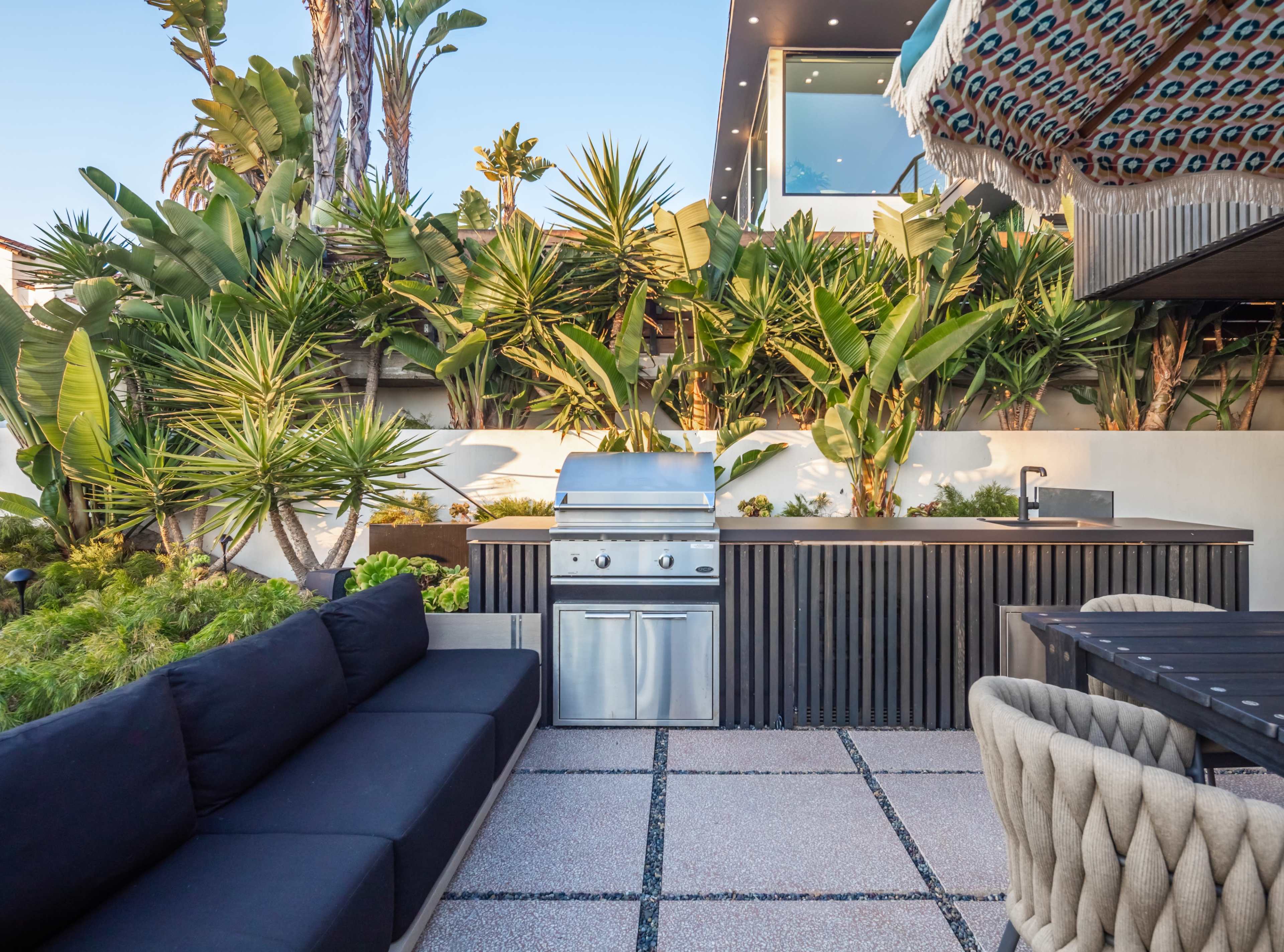 An outdoor kitchen area featuring a stainless steel grill, a dining table, and lush tropical plants surrounding a modern seating arrangement.