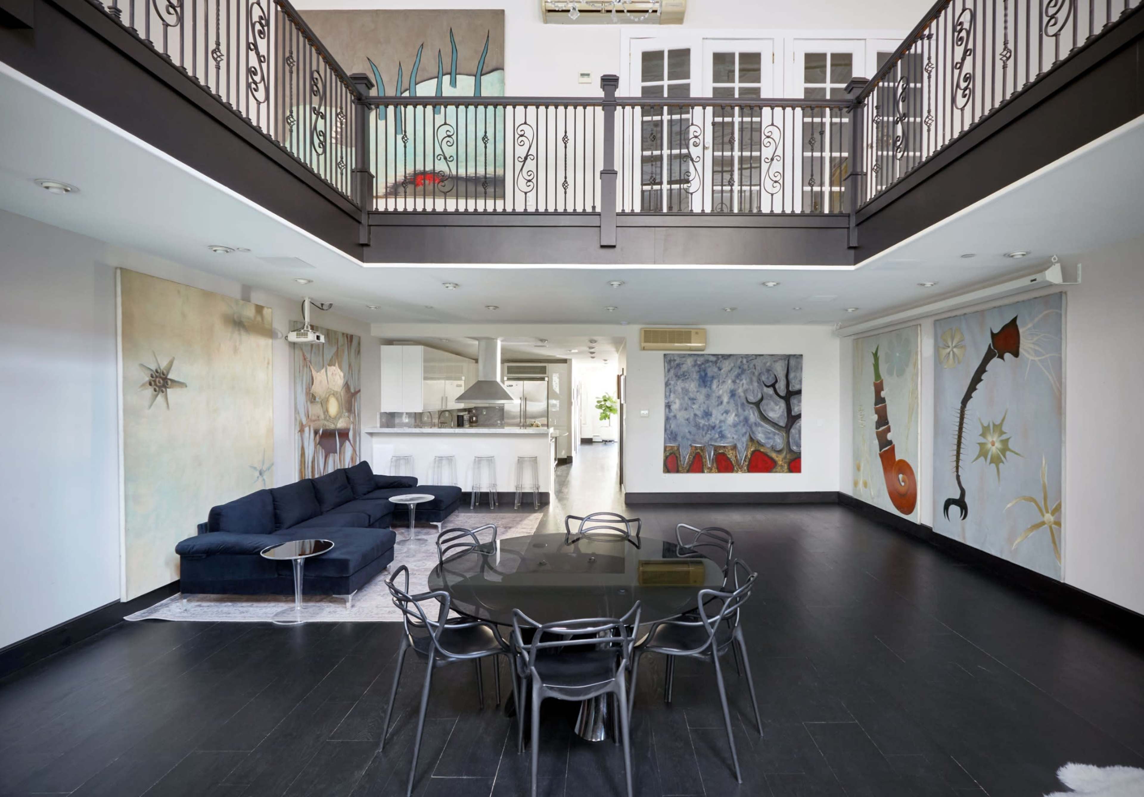 A modern, open-concept living space with a black dining table surrounded by transparent chairs and an upper balcony overlooking the kitchen area.