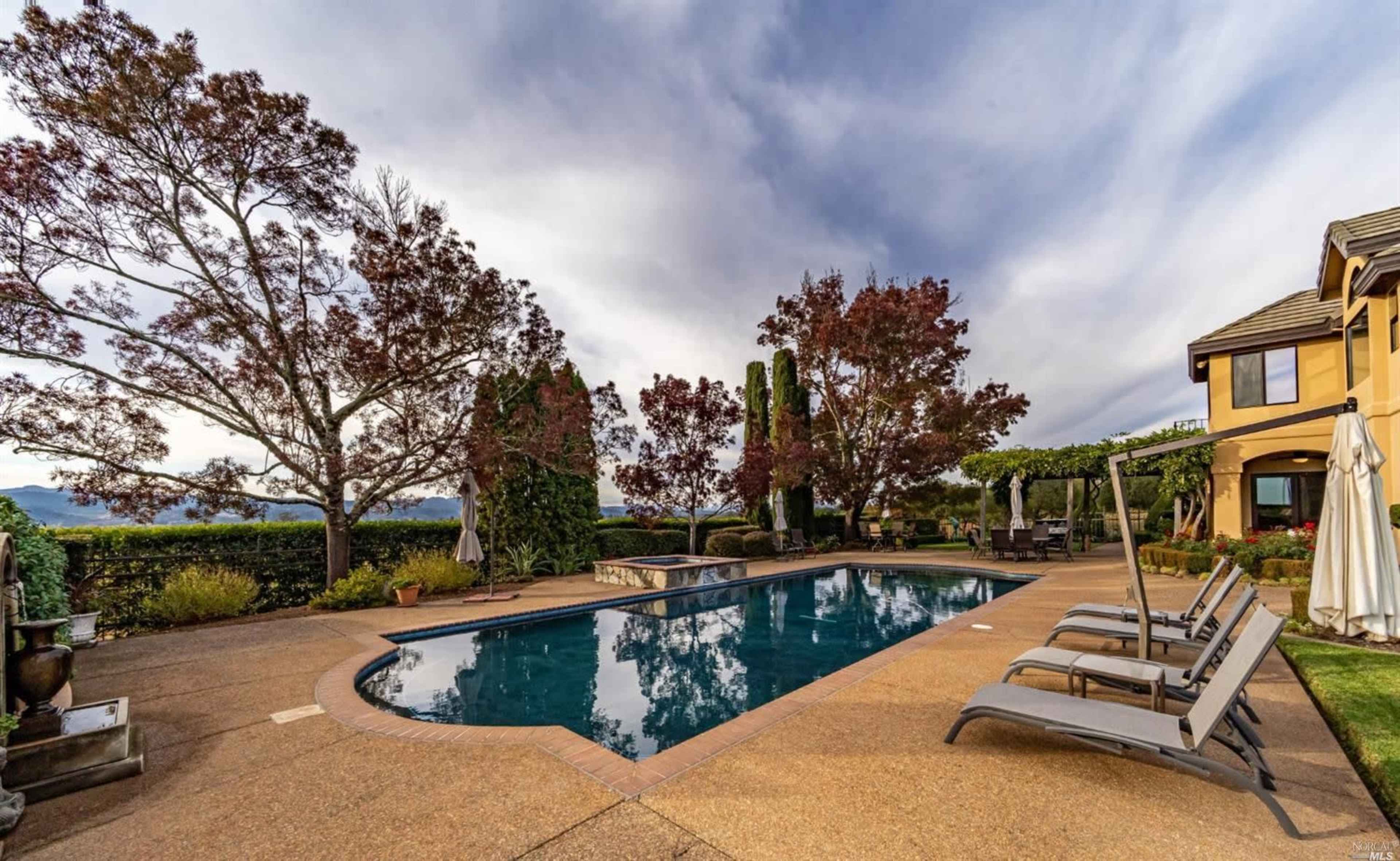 A rectangular swimming pool is bordered by lounge chairs and surrounded by landscaped gardens featuring trees and shrubs.