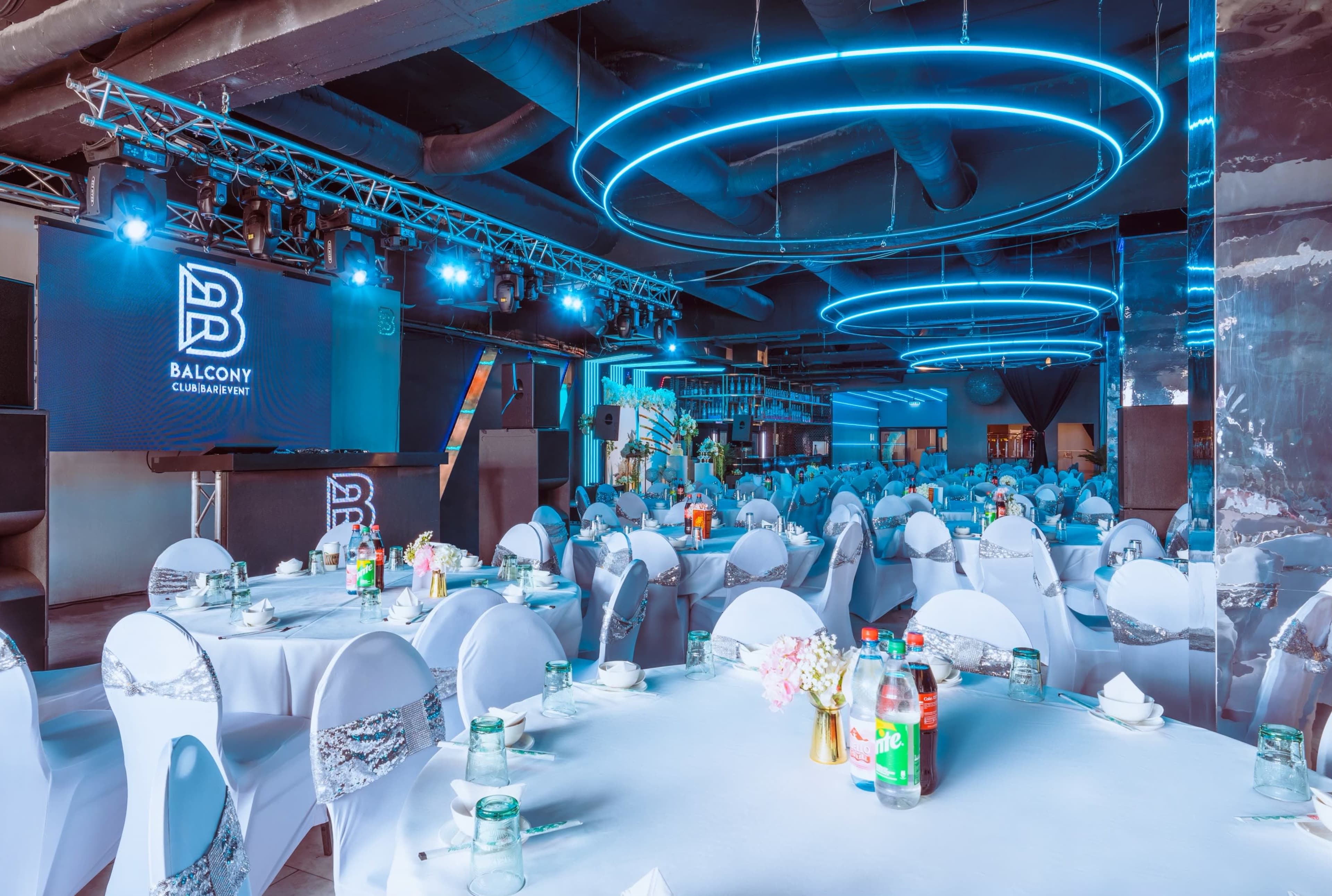 An elegantly arranged dining area with round tables covered in white tablecloths and chairs draped in white covers, illuminated by blue LED lights and featuring a stage in the background.