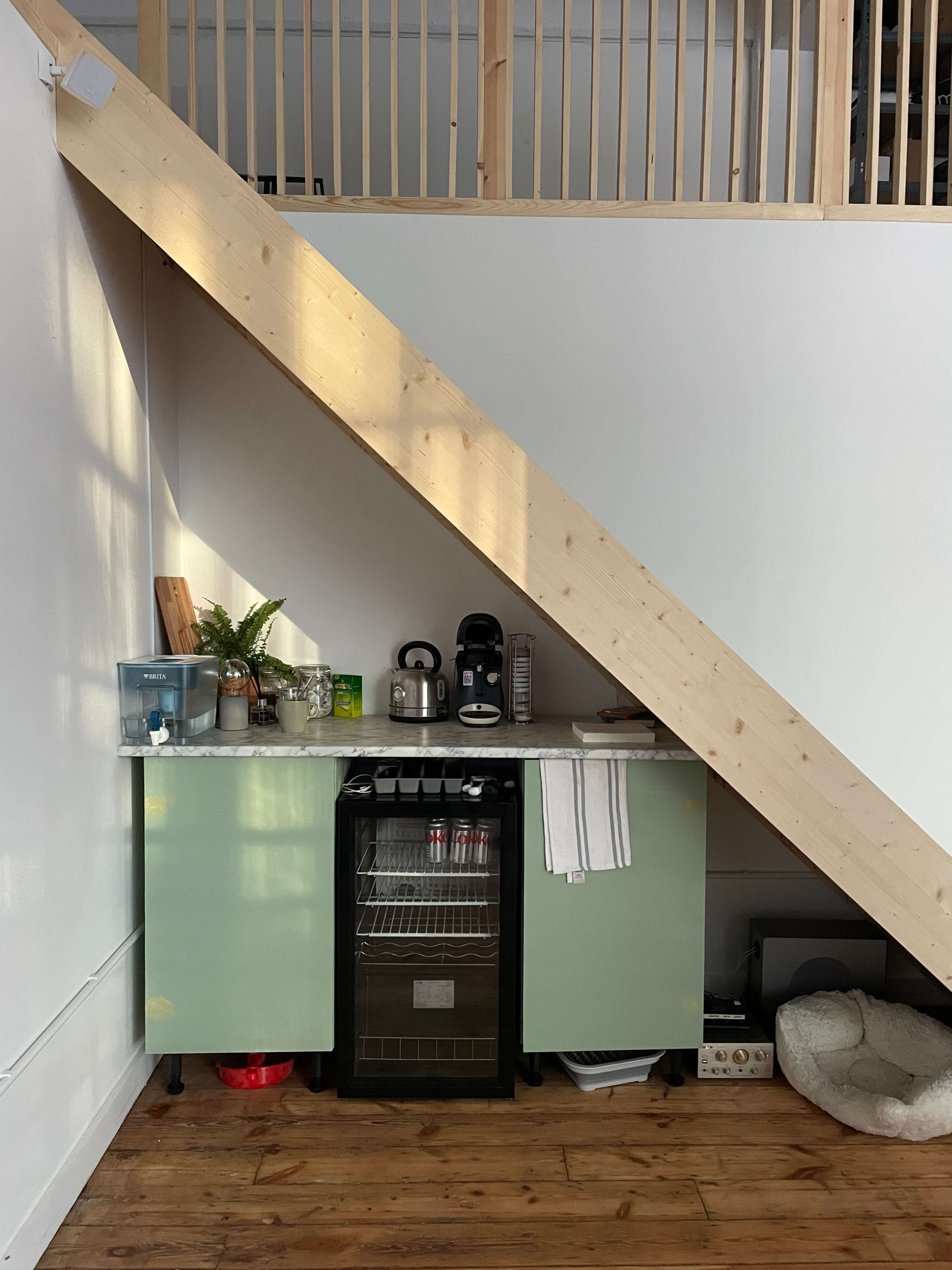 A compact kitchenette with a green cabinet, a mini-fridge, and a coffee maker is located under a wooden staircase.