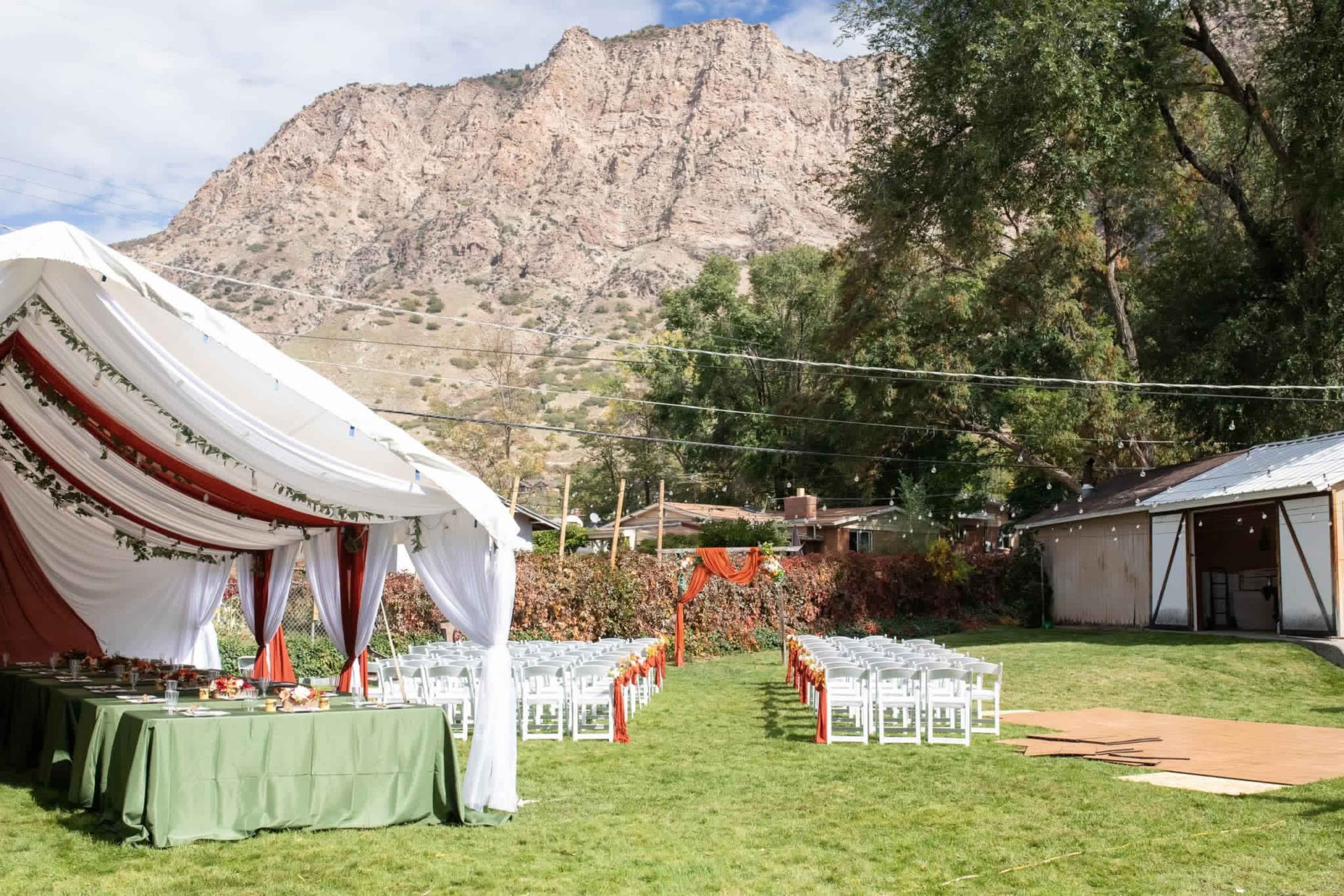 A tented area with tables is set up on a grassy field, facing rows of chairs arranged for an outdoor event against a backdrop of mountains.