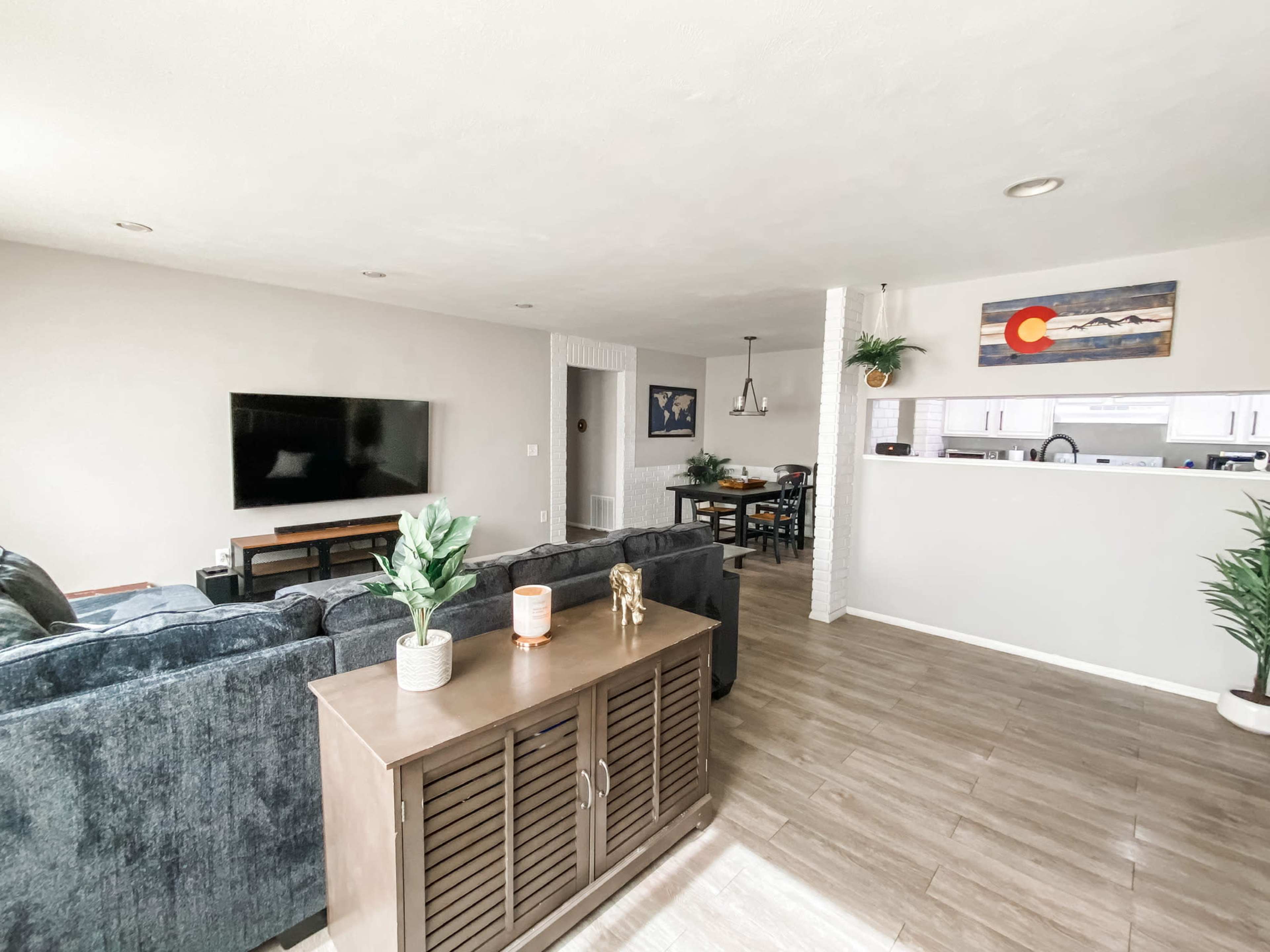 The living room features a dark couch facing a wall-mounted television, with a dining area visible in the background that includes a table and chairs.