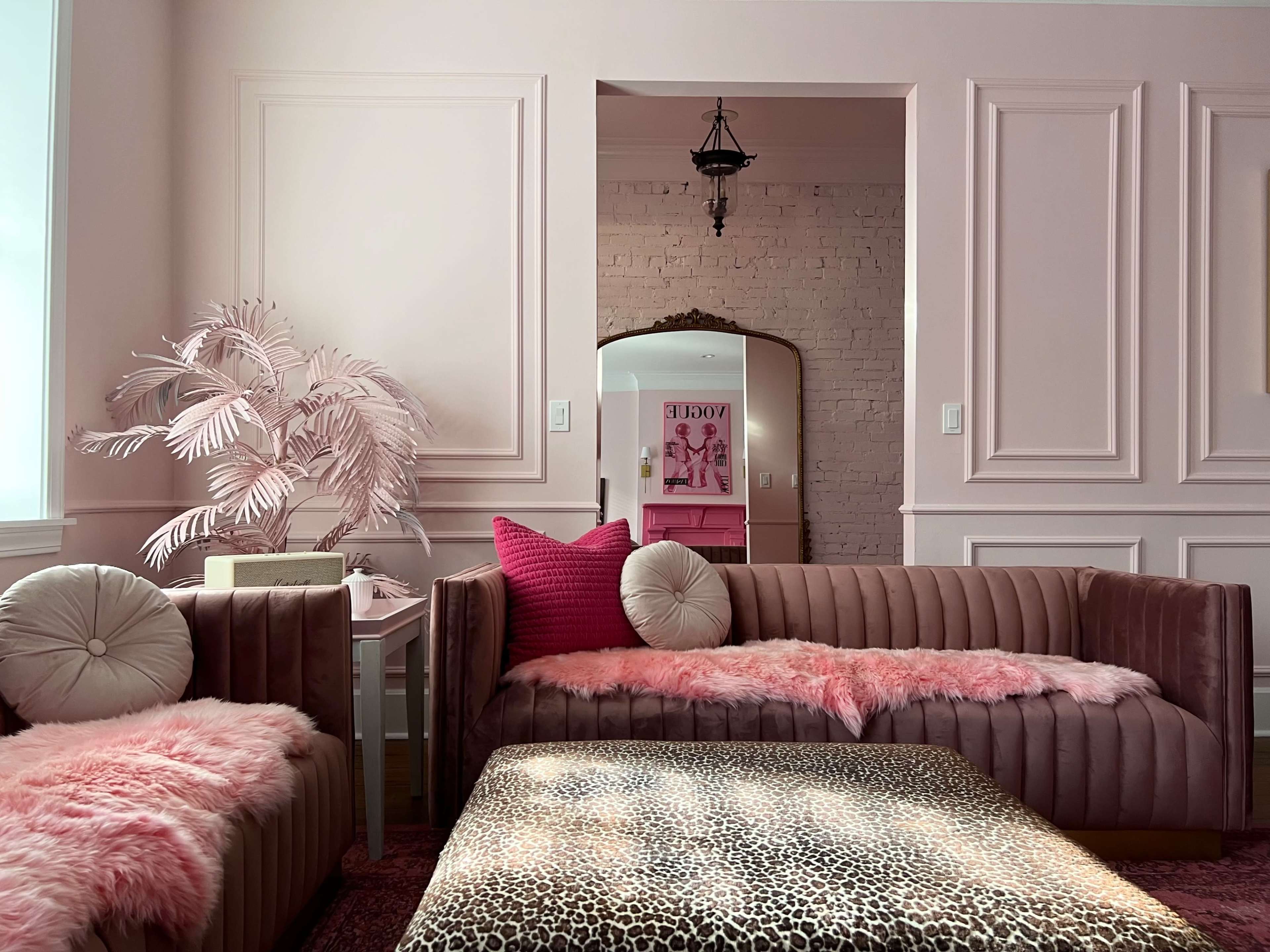 The room features two plush sofas draped with pink fur throws, a leopard-print coffee table, and a large mirror reflecting a stylish decor in soft pastel tones.