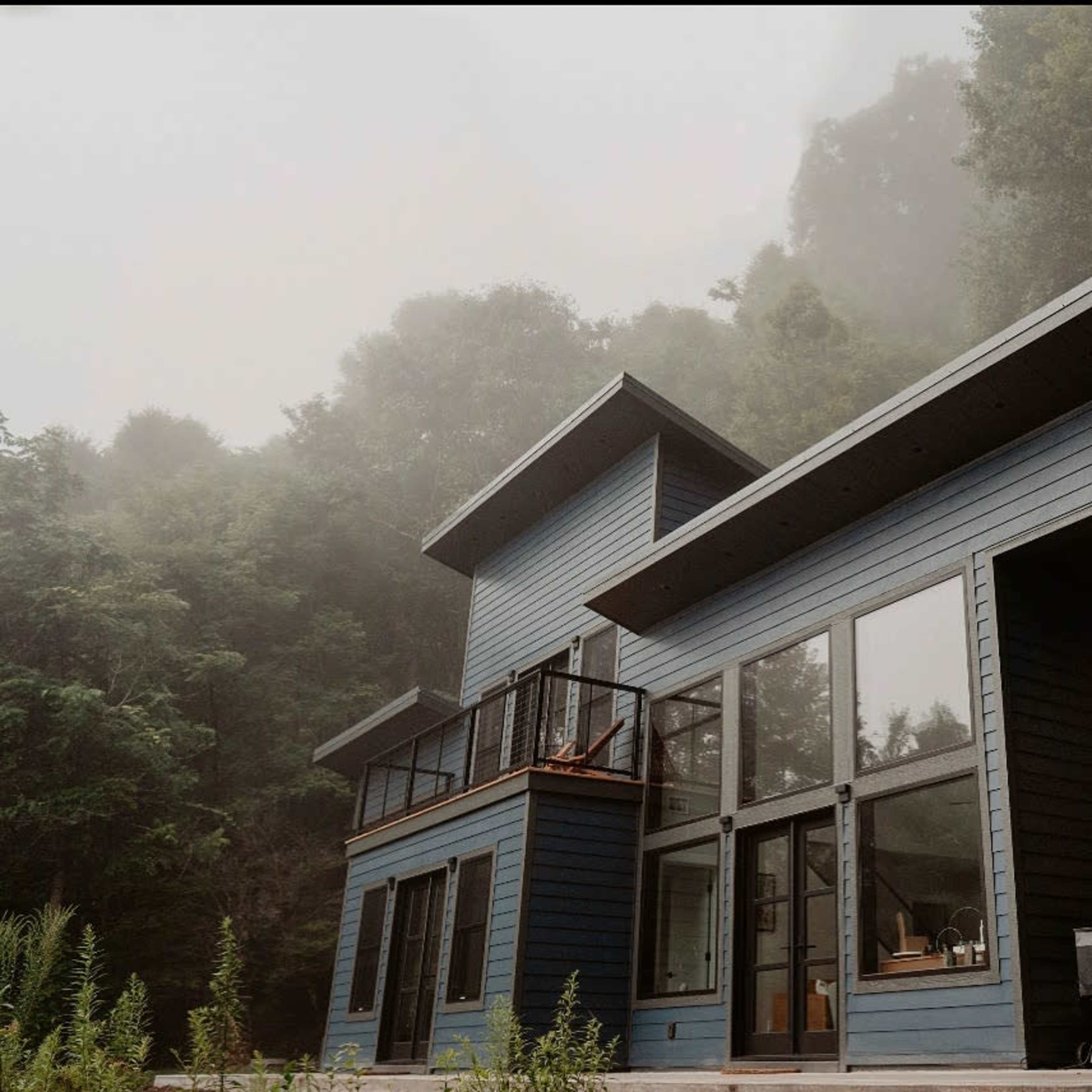 A modern blue house with large windows is situated in a foggy forested area.