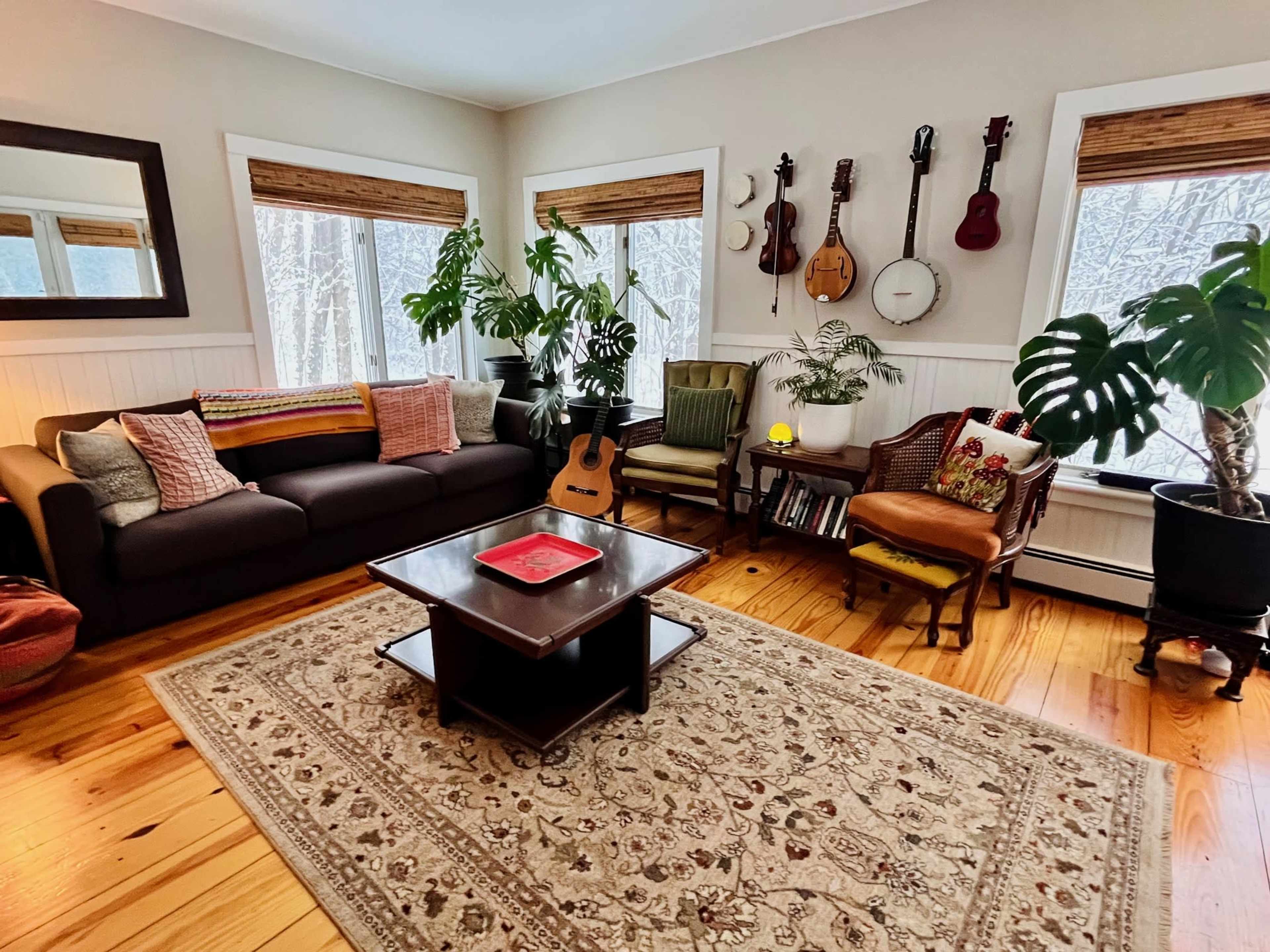 The living room features a dark sofa, a coffee table, various musical instruments on the wall, and large plants, all set against a backdrop of large windows.