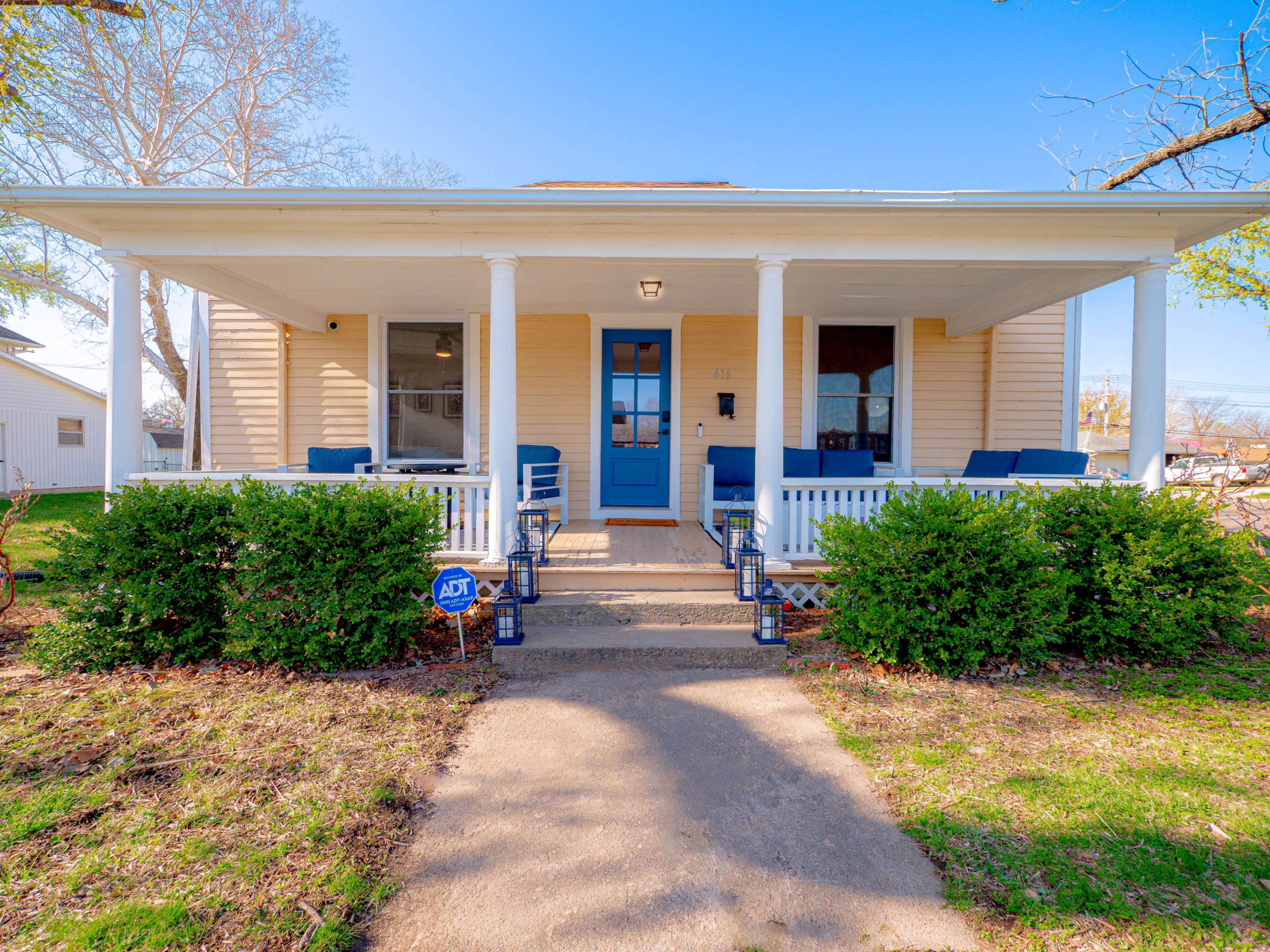 The image shows a yellow, single-story house with a front porch, a blue door, and two sets of chairs on either side of the entrance.