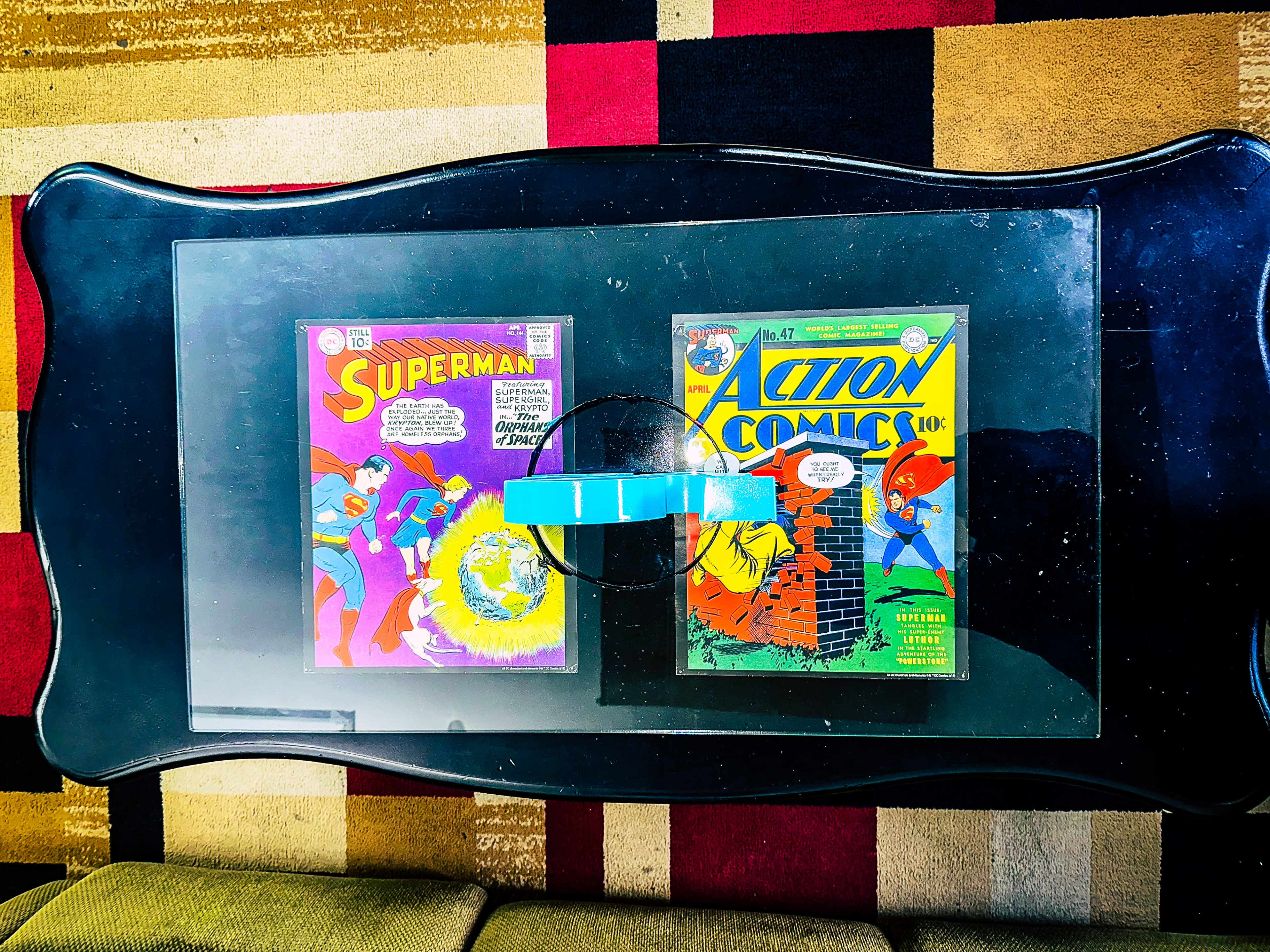 Two comic books, "Superman" and "Action Comics," displayed on a glass coffee table with a turquoise coaster in between them.