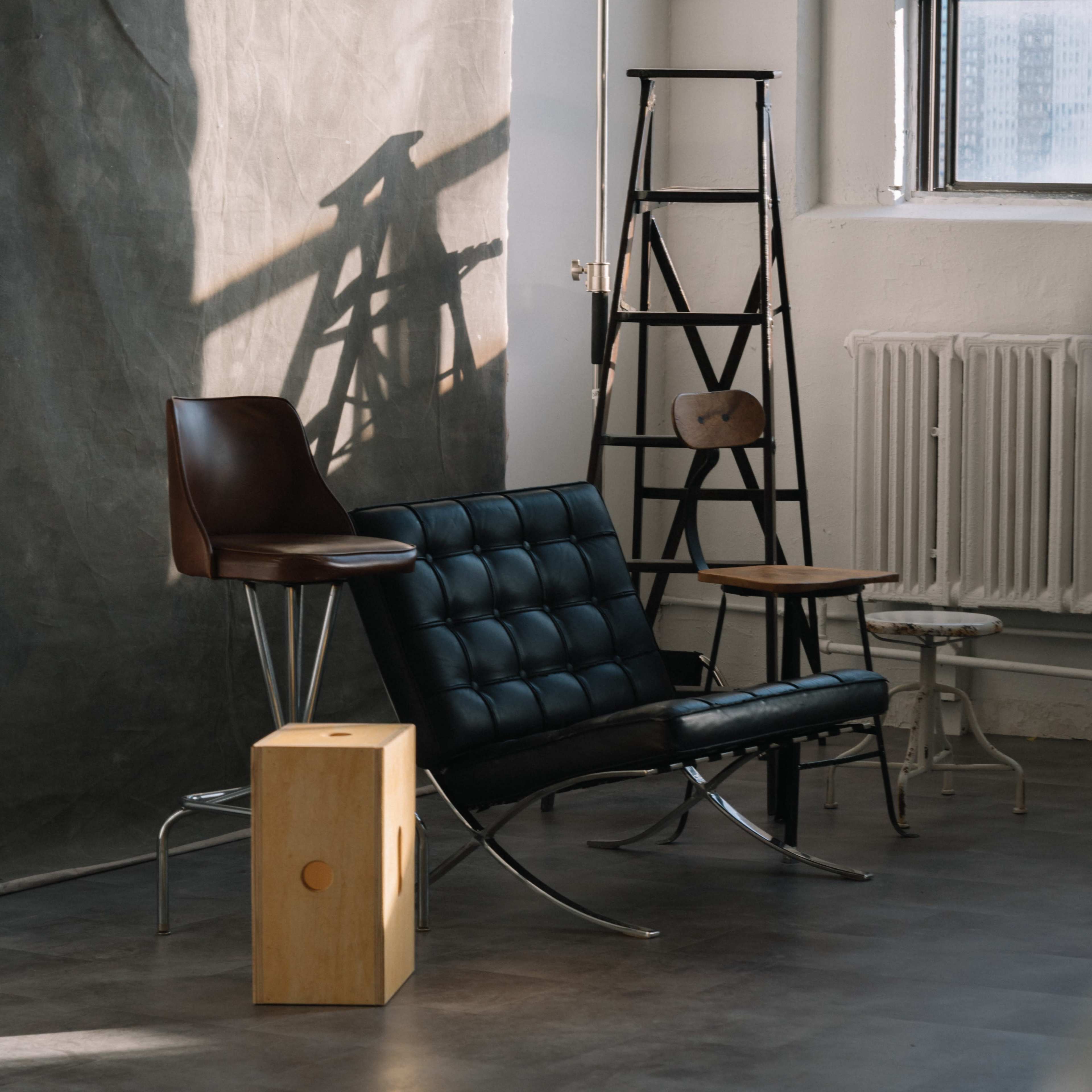 The scene features a variety of chairs, including a modern black lounge chair, a wooden stool, and a metal ladder, arranged near a window with natural light casting shadows on the wall.