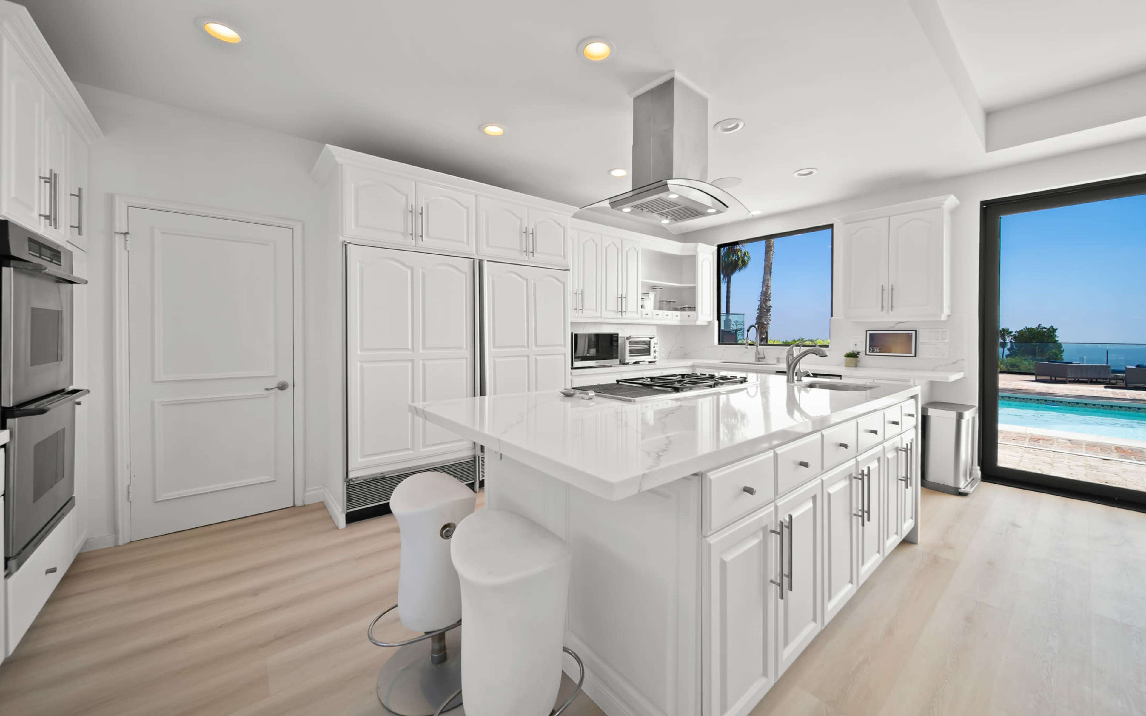 A modern kitchen features white cabinetry, a central island with a gas stove, and large windows that overlook a pool and ocean view.