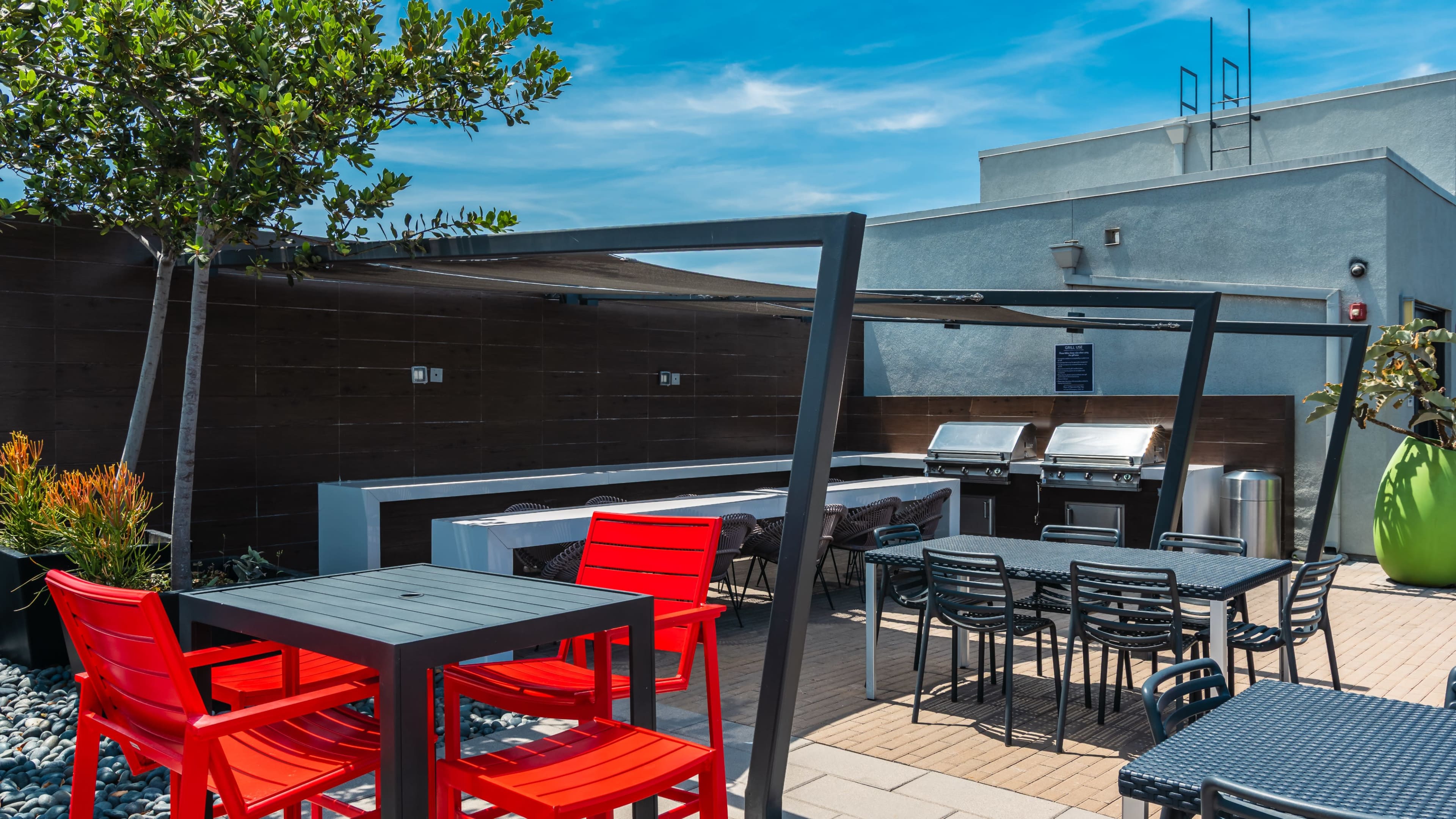 The image shows an outdoor patio area featuring red chairs and tables, a grill station under a canopy, and green landscaping against a blue sky.