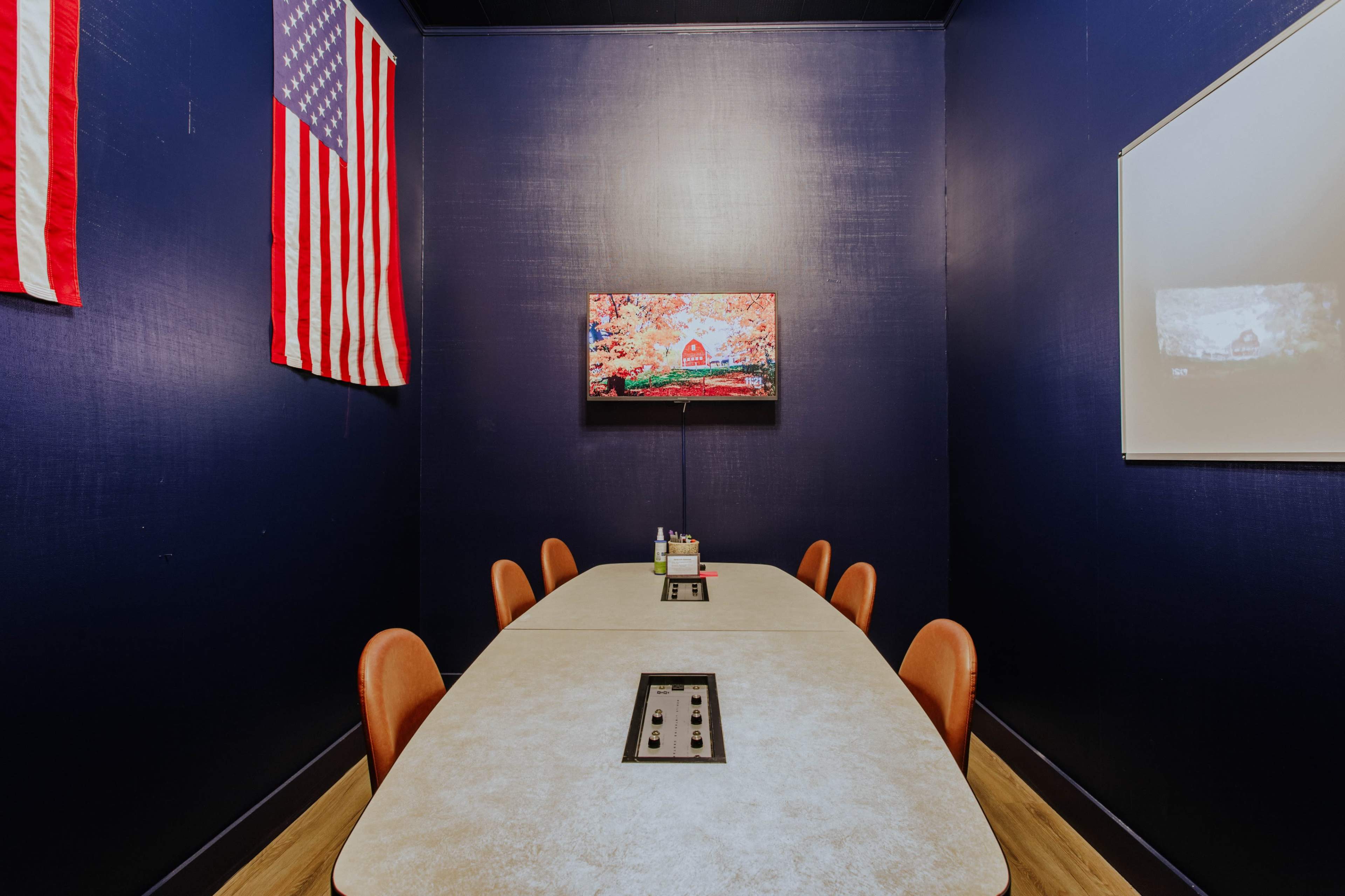 A conference room features a long table surrounded by orange chairs, with American flags on the walls and a TV mounted above the table.