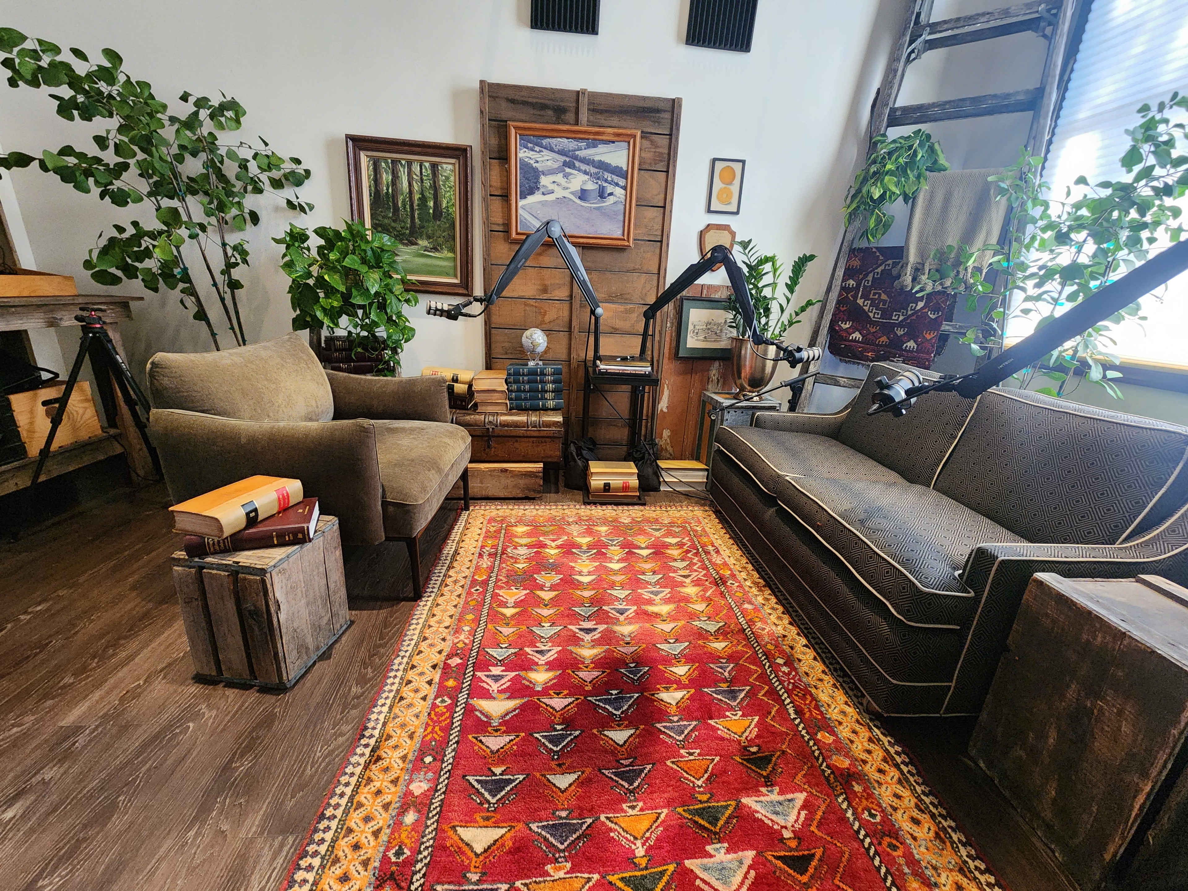 The image shows a cozy sitting area with two sofas, a patterned rug, wooden crates, and potted plants, surrounded by artwork on the walls and a wooden ladder.