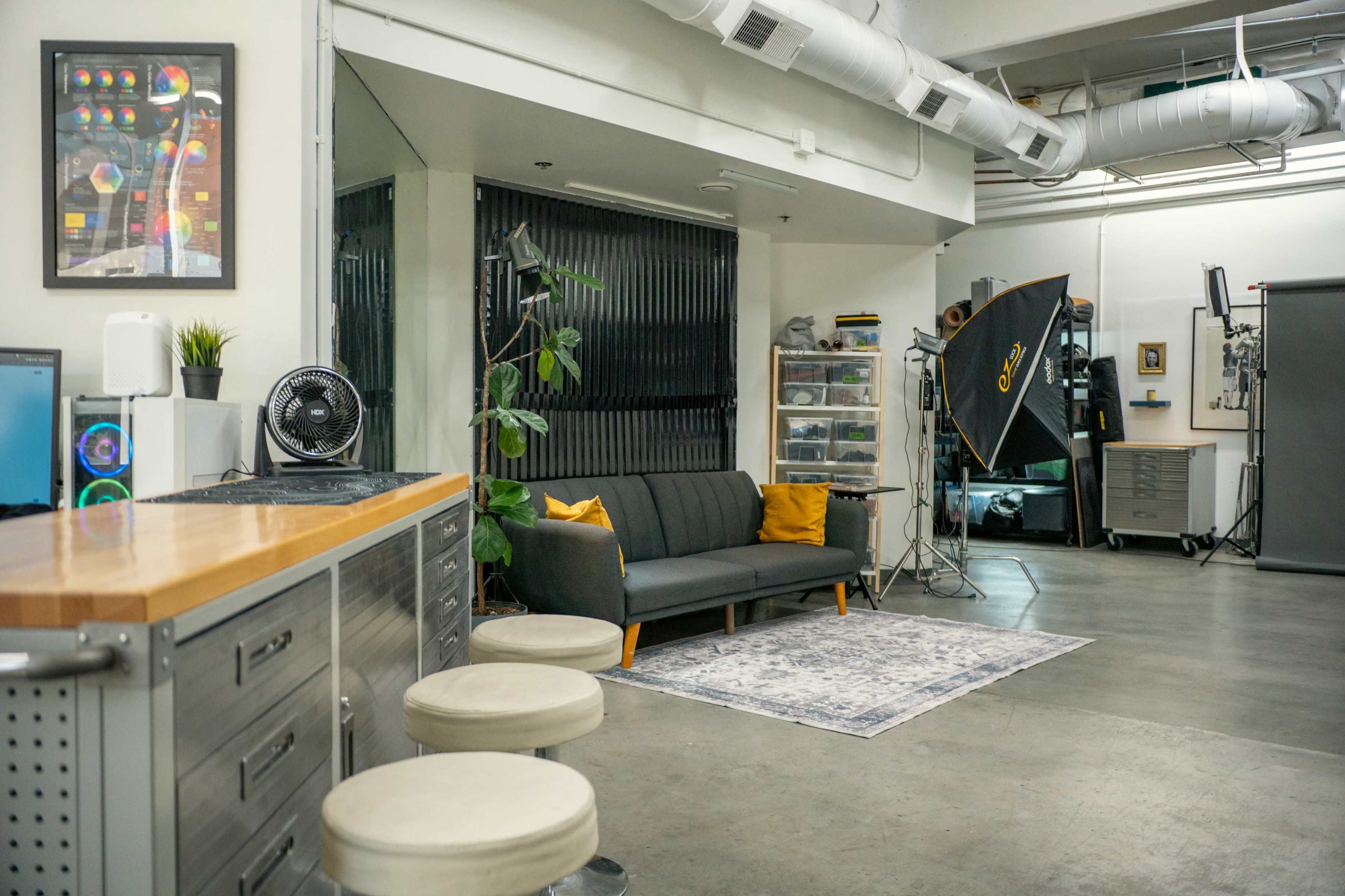 The image shows a modern studio space featuring a seating area with a sofa and plants, photography equipment, and a metal workbench.