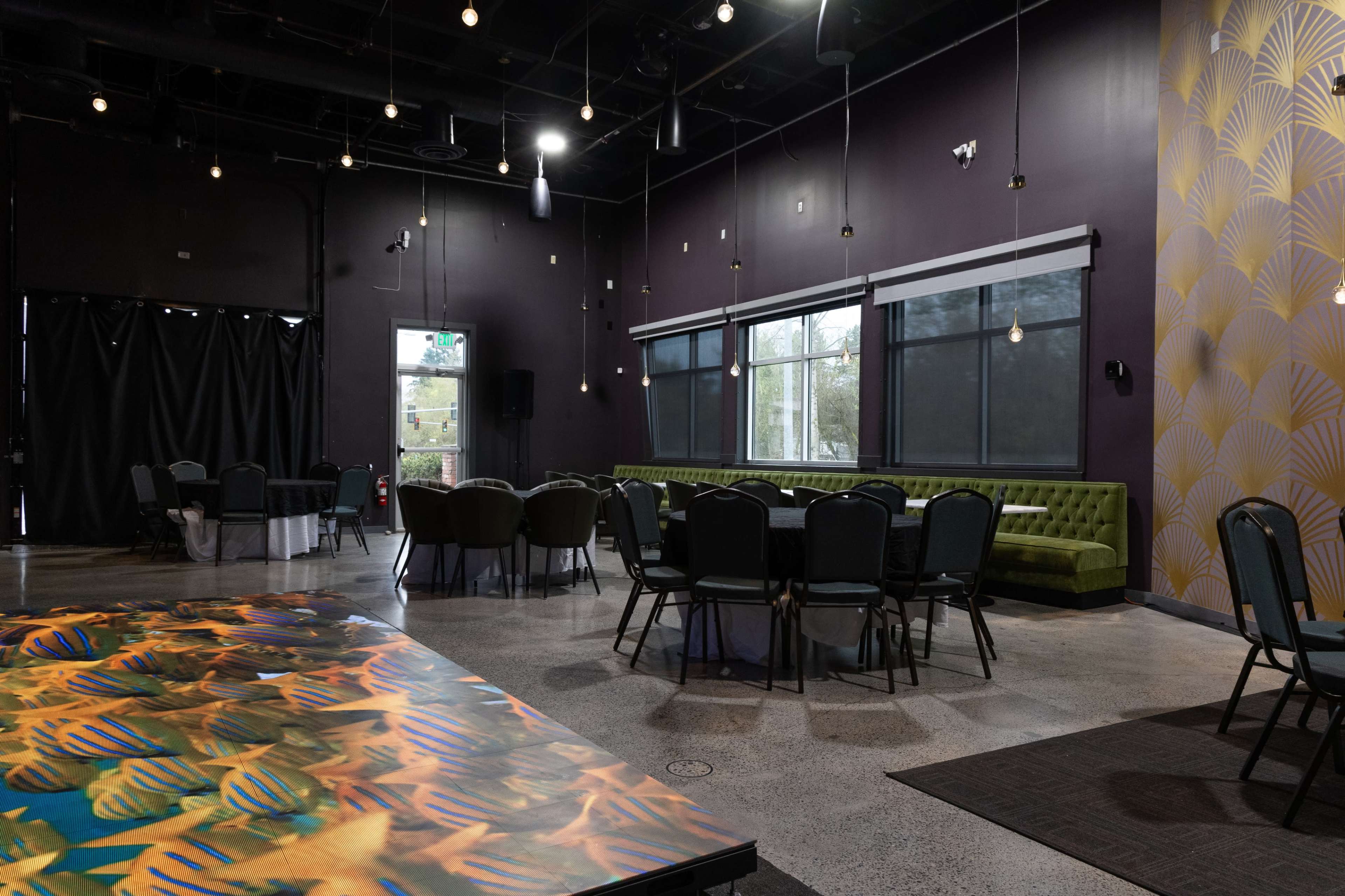 The image shows a modern event space with a green couch, round tables, and large windows, illuminated by hanging light bulbs.