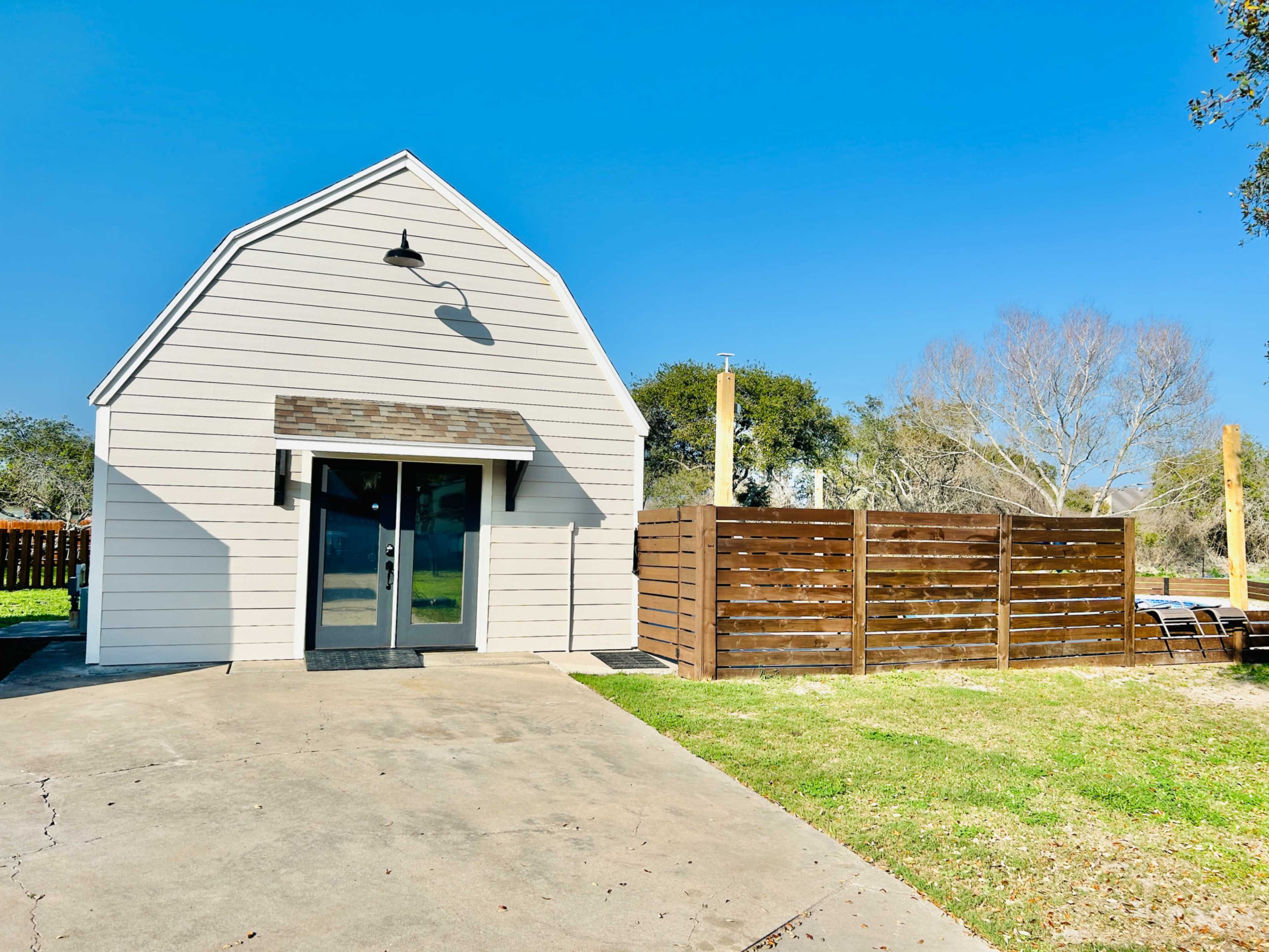 A small, gabled house with a paved driveway is situated beside a fenced yard against a clear blue sky.