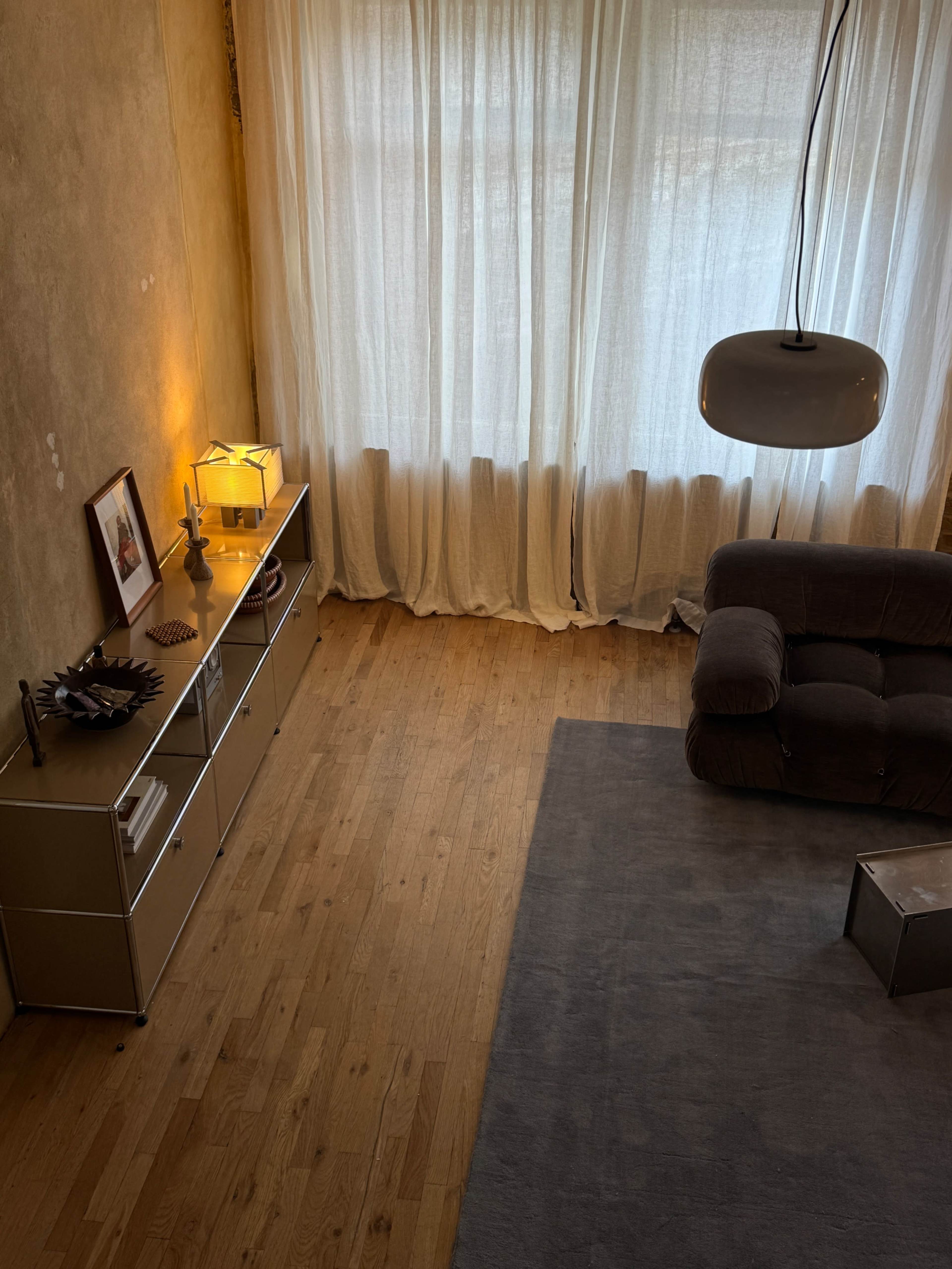 The image shows a spacious room with wooden flooring, a gray area rug, a brown couch, a console table with decorative items, and large windows covered with sheer curtains.