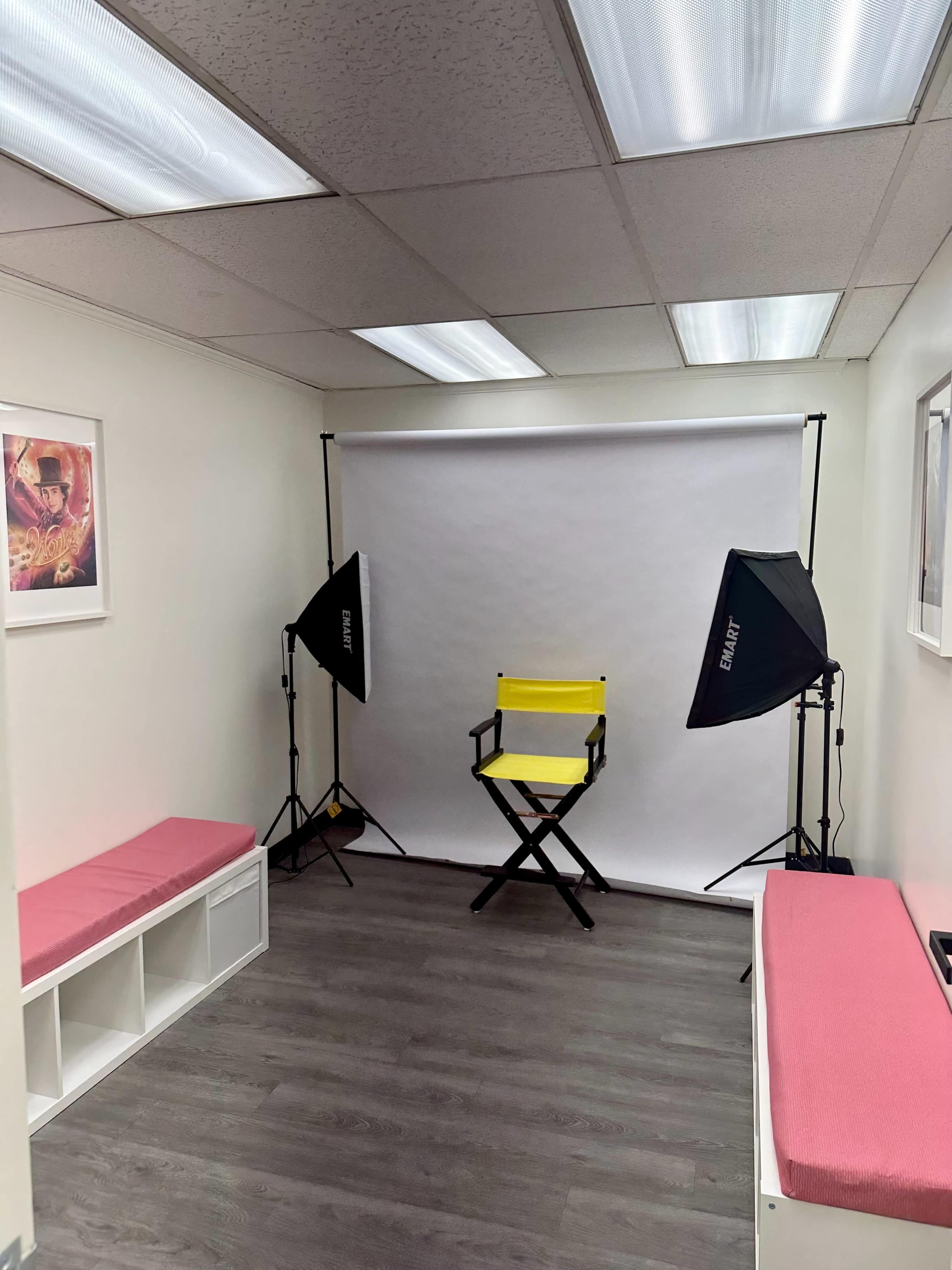 The image shows a simple studio space with a yellow director's chair positioned in front of a white backdrop, flanked by two lighting setups, and surrounded by pink benches and framed artwork.