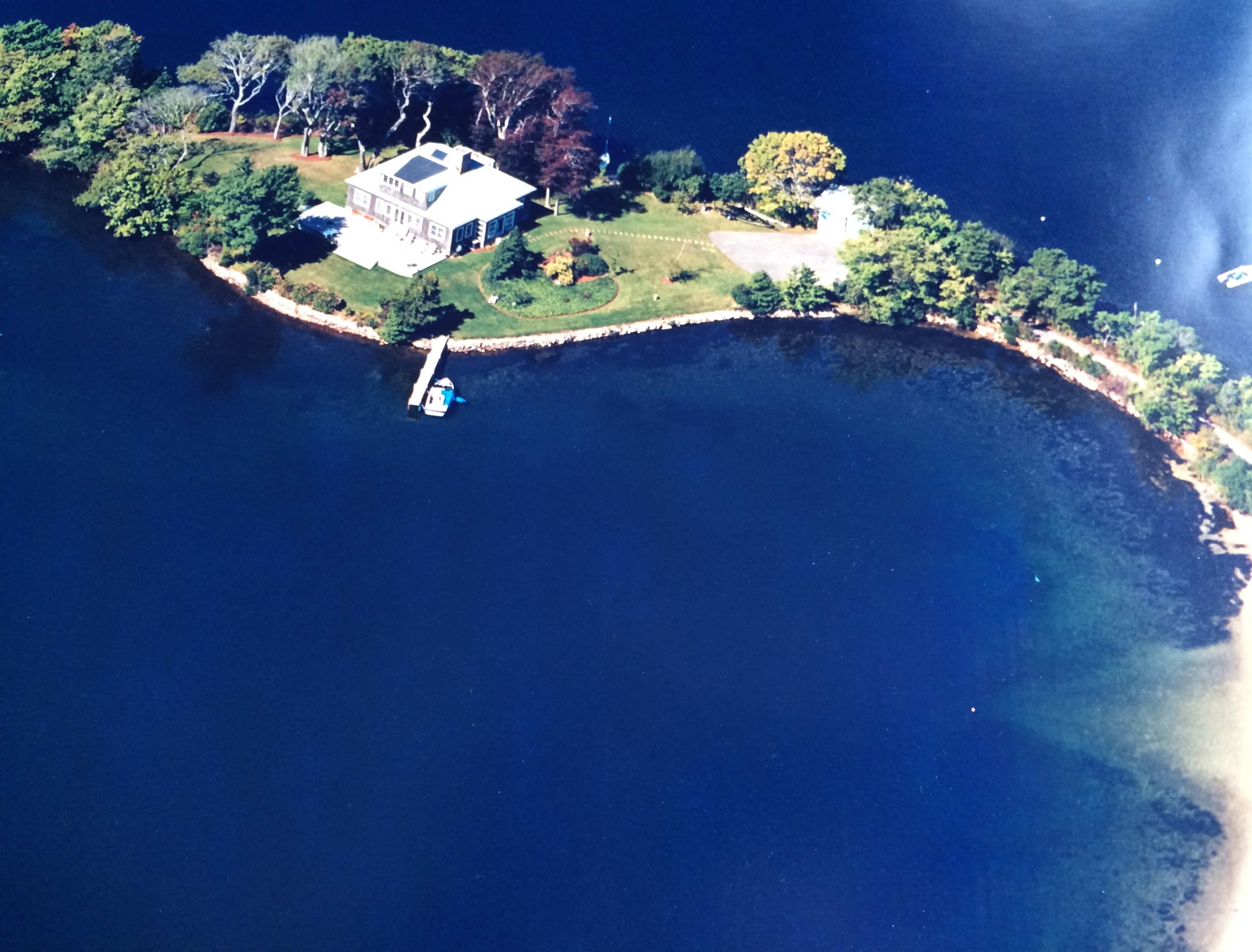 An aerial view shows a house situated along a curved shoreline with a dock and surrounding greenery.