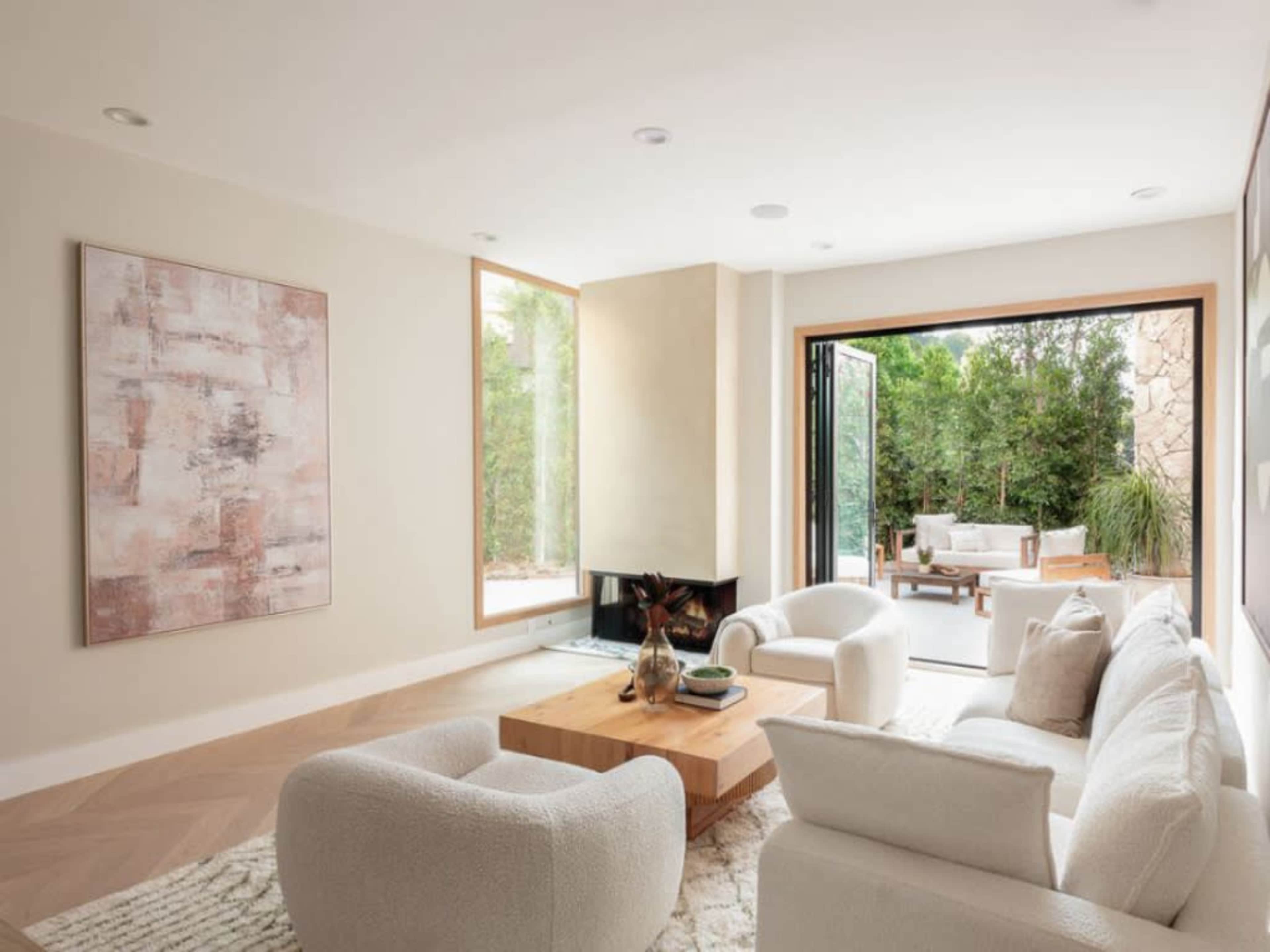 A modern living room features a light-colored sofa and armchairs arranged around a wooden coffee table, with large windows opening to a patio and greenery outside.