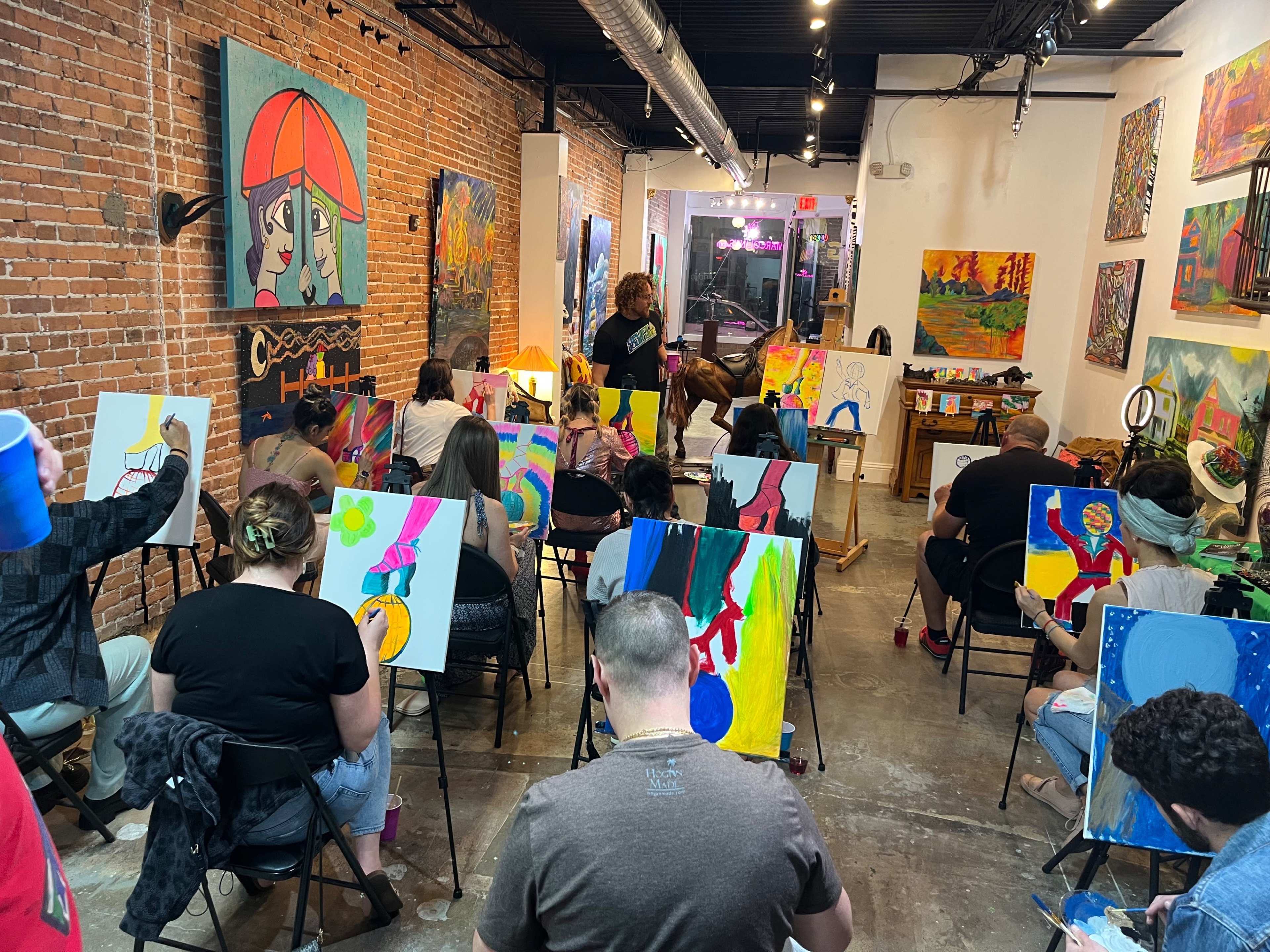 A group of people participate in a painting class in an art studio with exposed brick walls and various colorful artworks displayed.