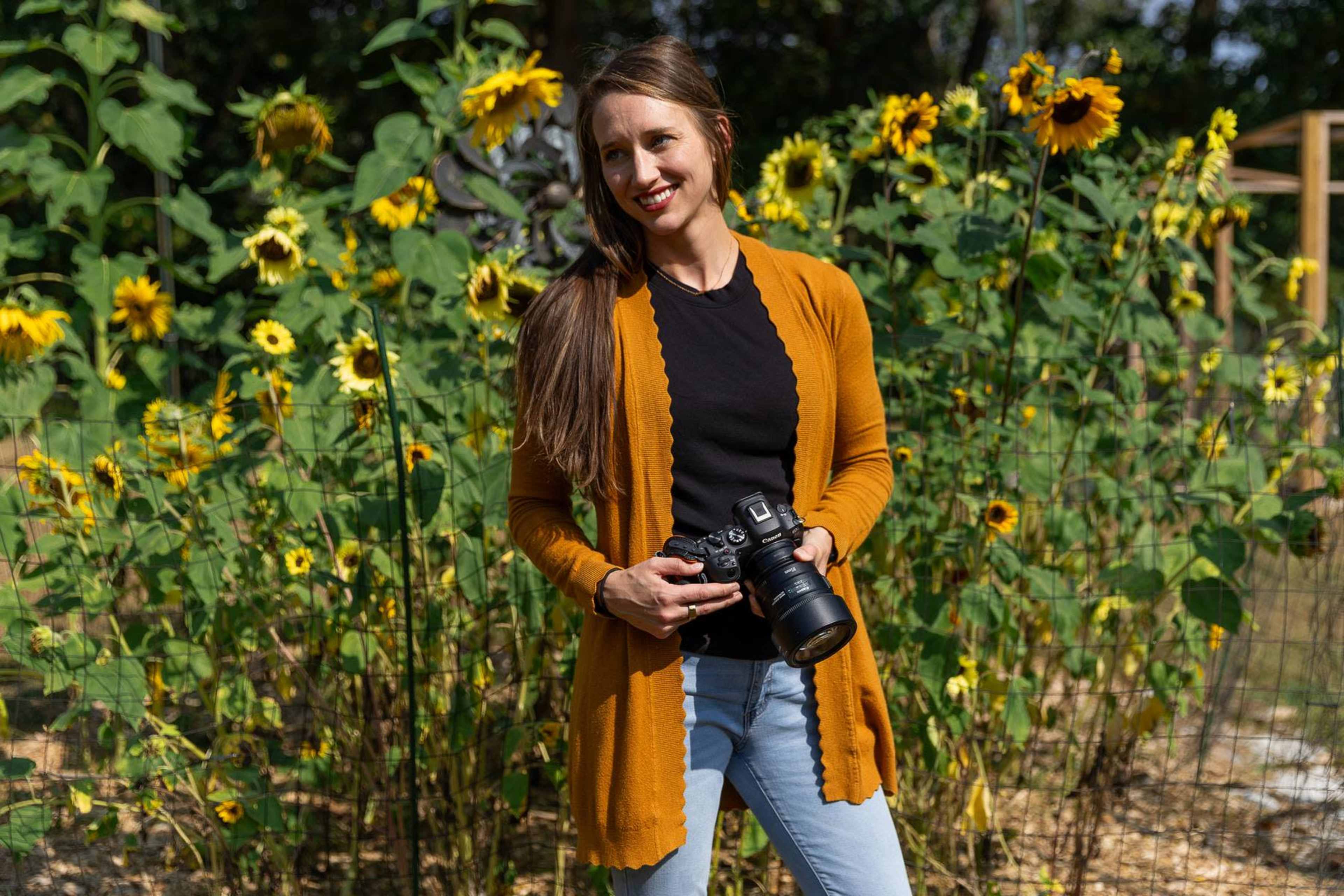 A woman holds a camera while standing in front of a field of sunflowers.