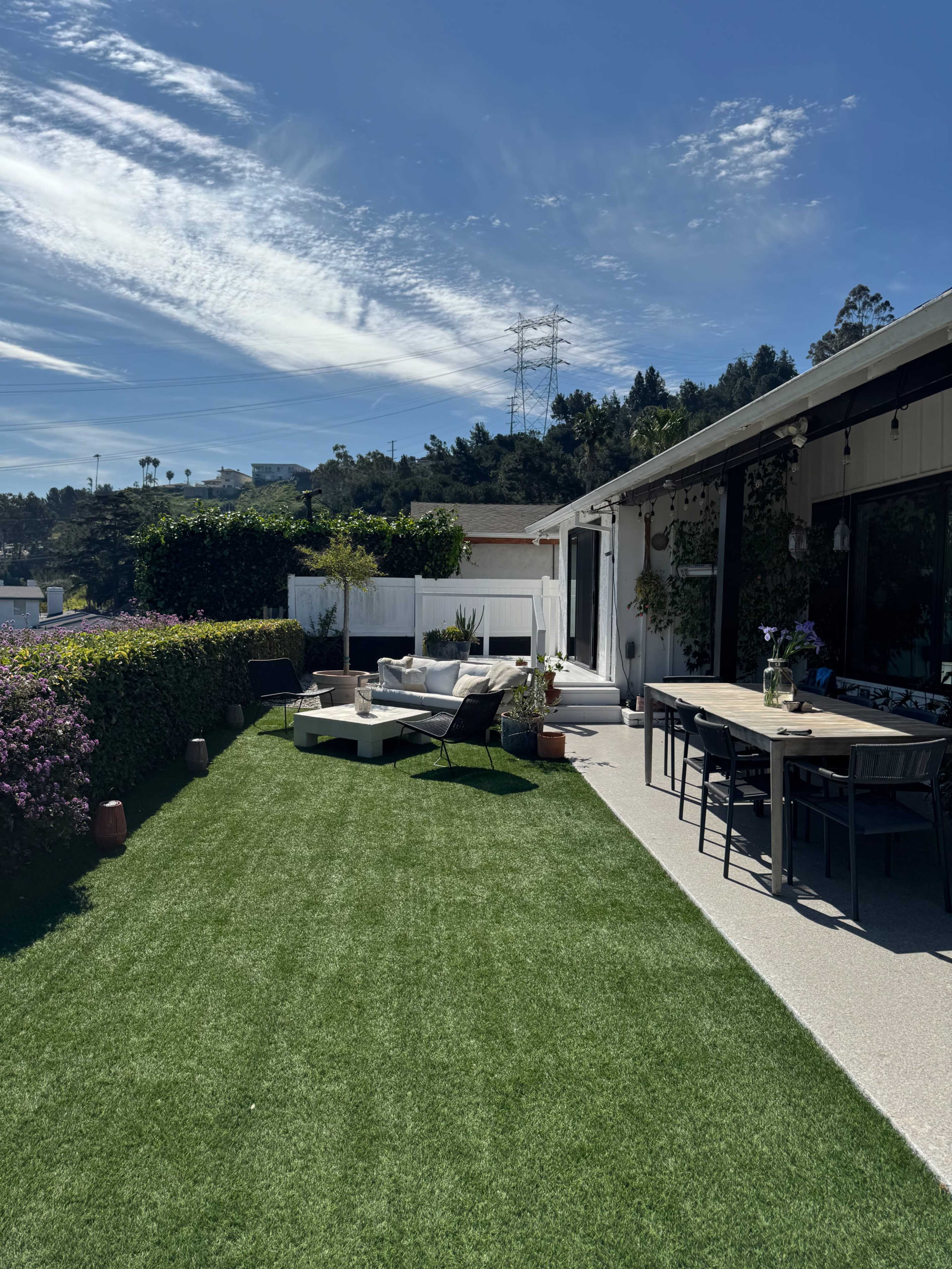 The image shows a landscaped outdoor patio area with artificial grass, a seating arrangement, and a dining table overlooking a hillside.