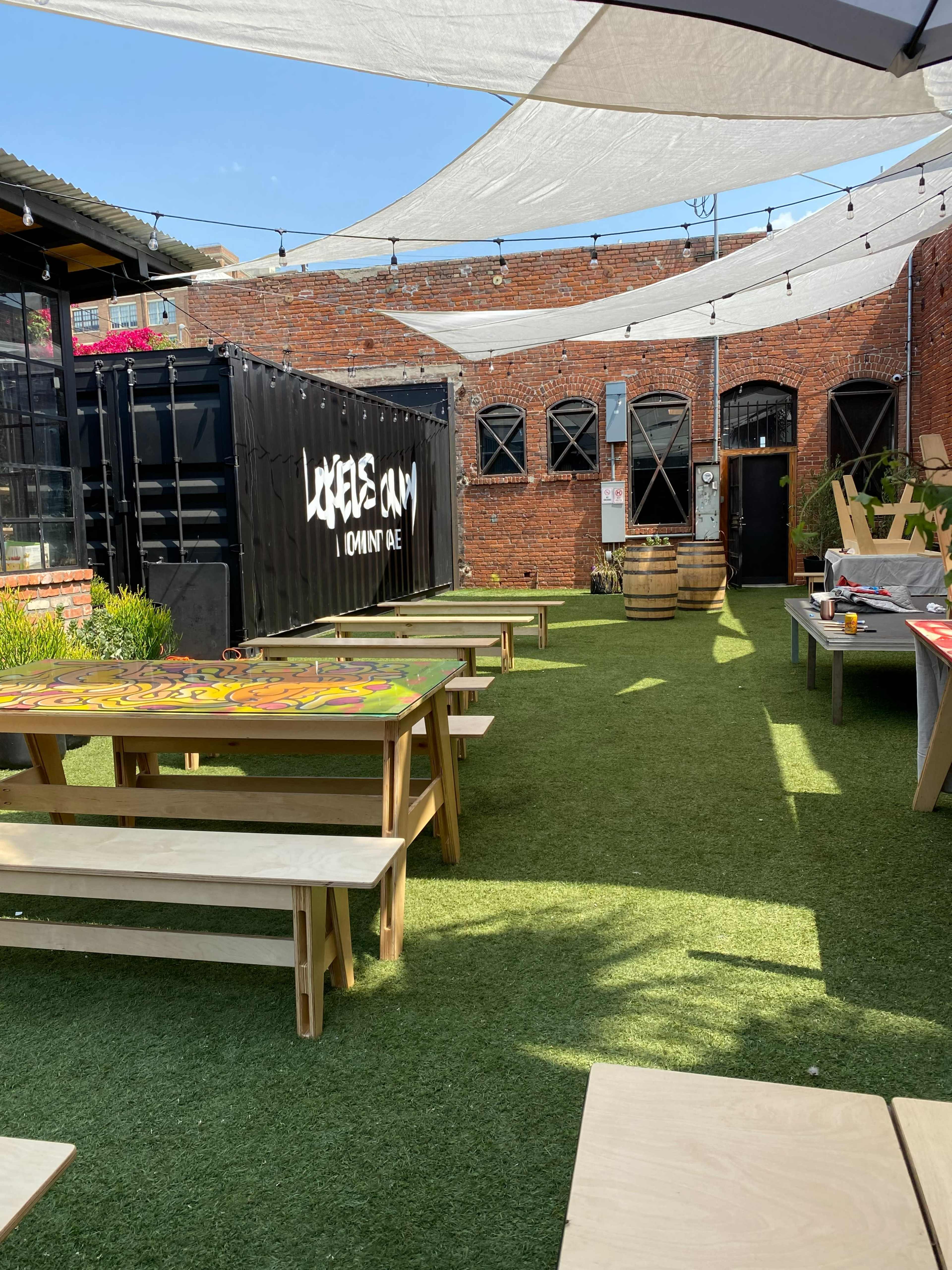 An outdoor area with green turf, wooden tables, and a shipping container, all enclosed by brick walls and shaded by fabric canopies.