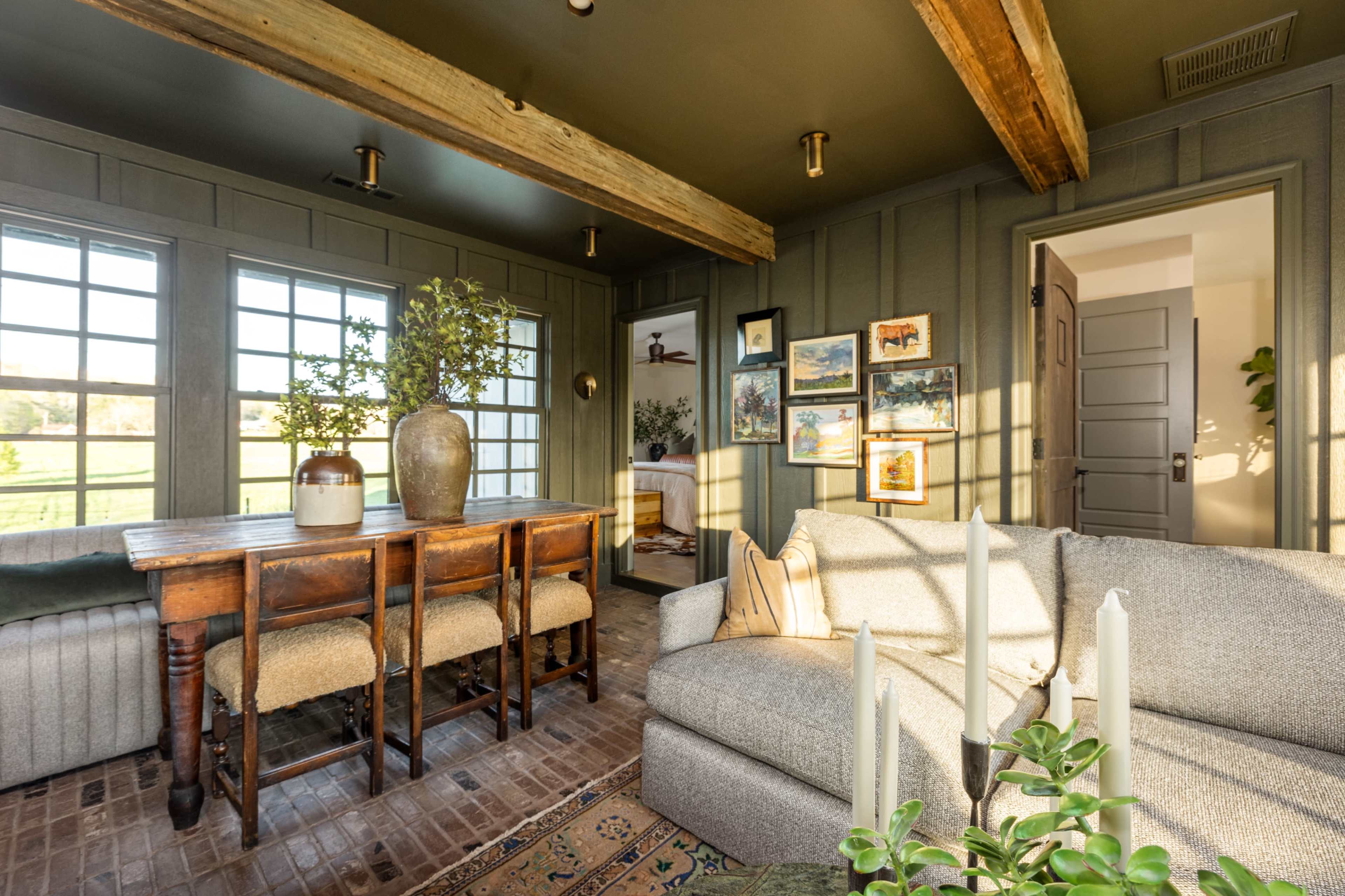 A cozy room with a wooden table, a gray sofa, and windows that provide a view of the outdoors, decorated with plants and framed pictures on the walls.