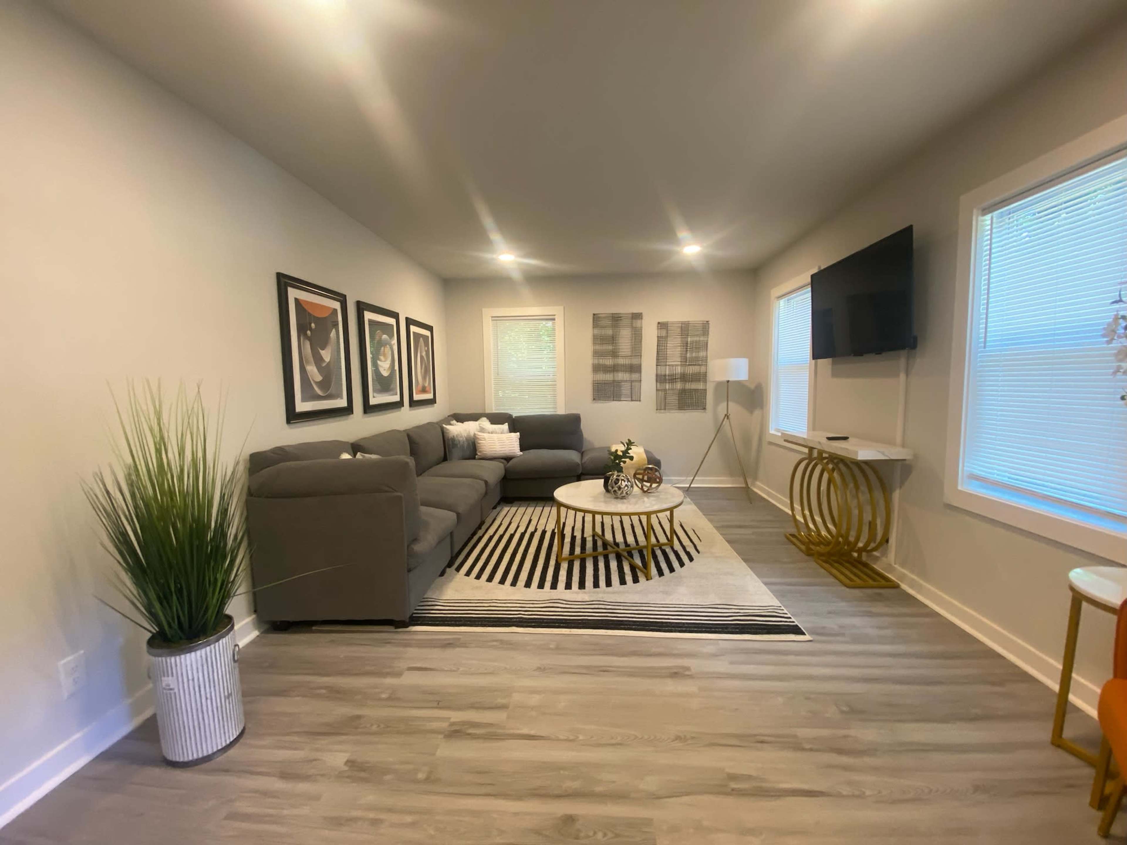 The living room features a gray sectional sofa, a circular coffee table on a striped rug, and a wall-mounted TV, along with neatly arranged decorative art and plants.