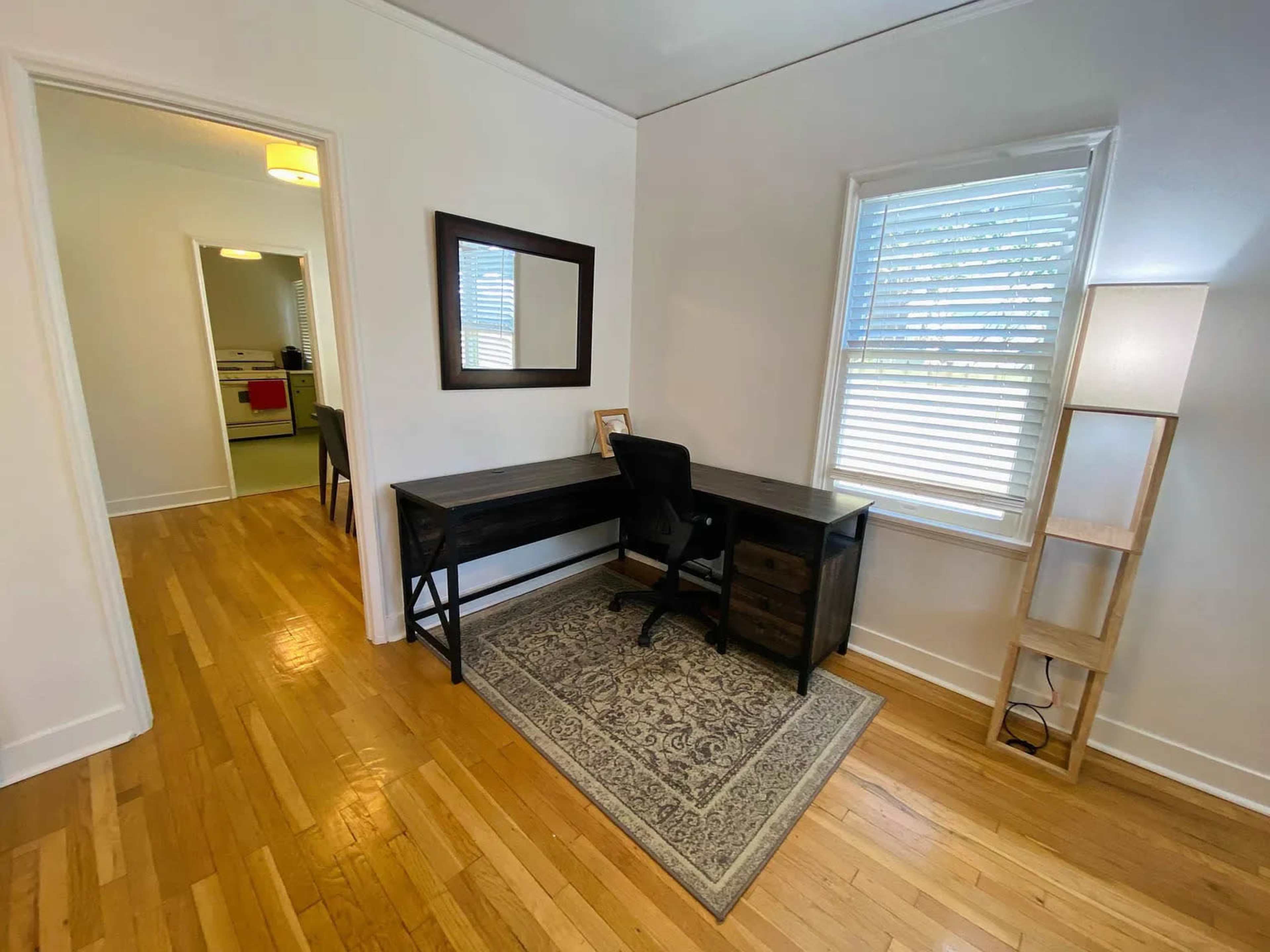 A small office space features a dark wooden desk with a chair, a mirror on the wall, and a floor lamp beside a window, with a view of an adjacent room.