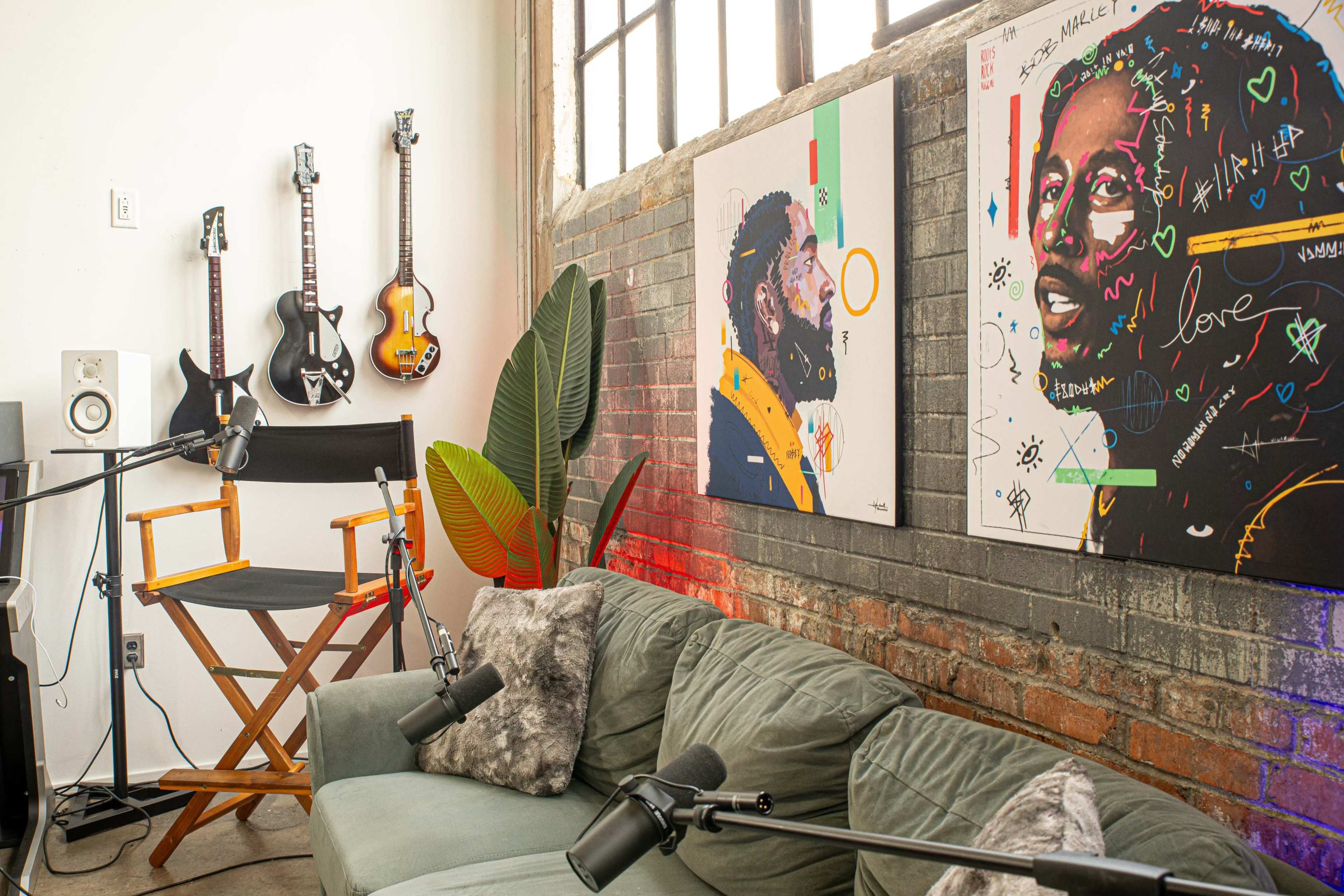 A cozy recording studio with two colorful portraits on the wall, guitars hanging, a microphone setup, and a green couch.