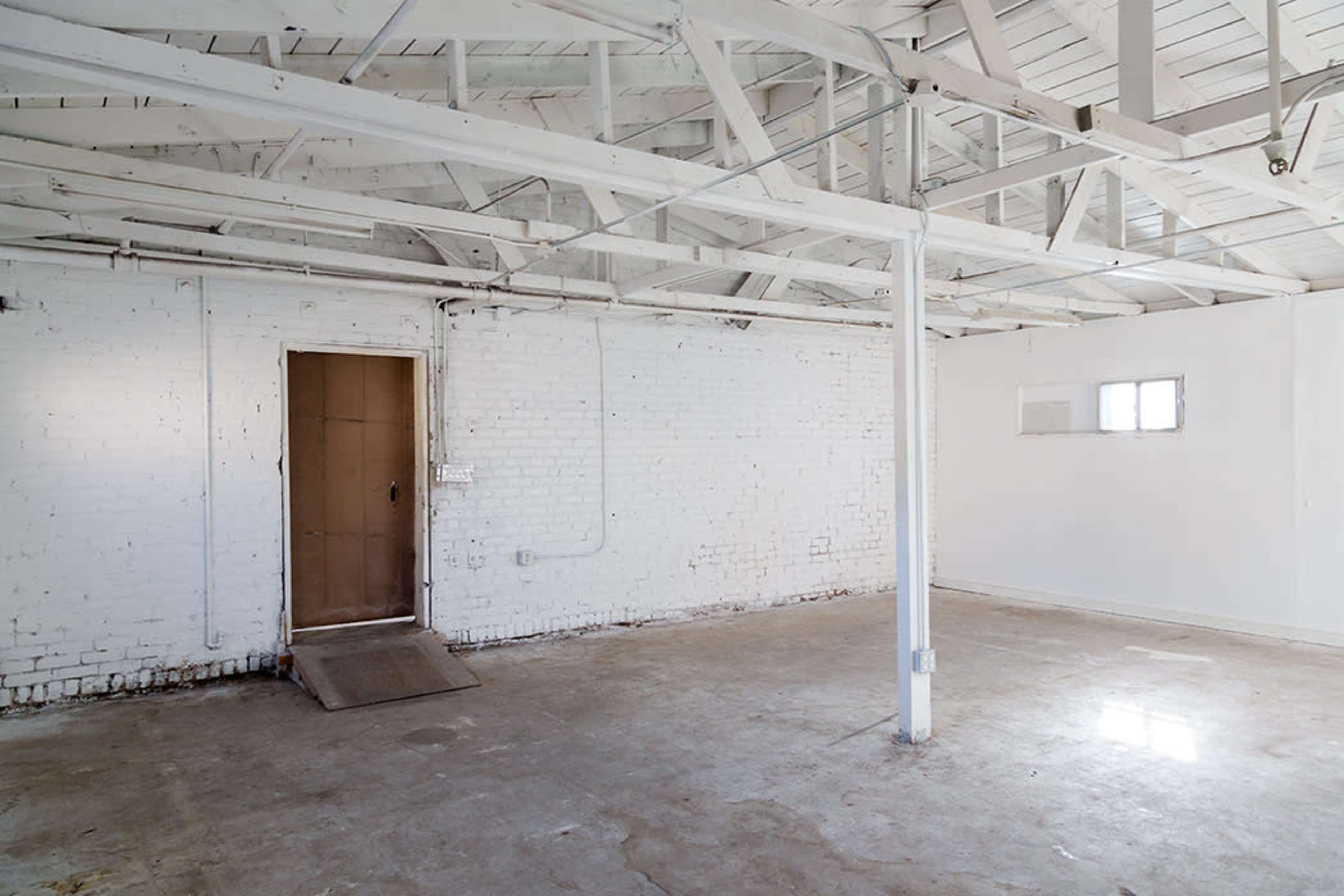The image shows a spacious, empty room with white brick walls, a concrete floor, and a door leading to the outside.
