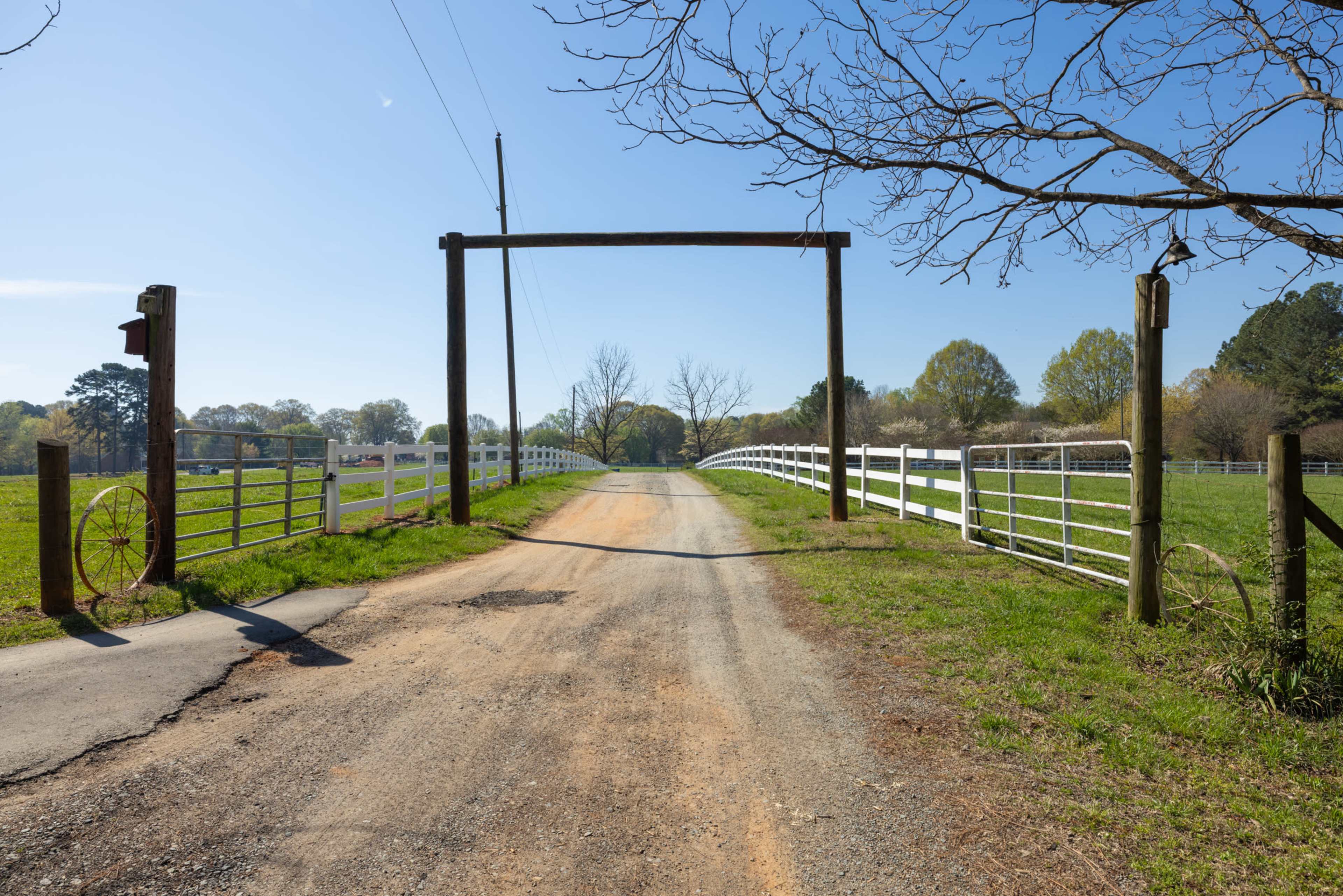 A gravel road leads through a wooden gate entrance, flanked by white fences and trees in the background.