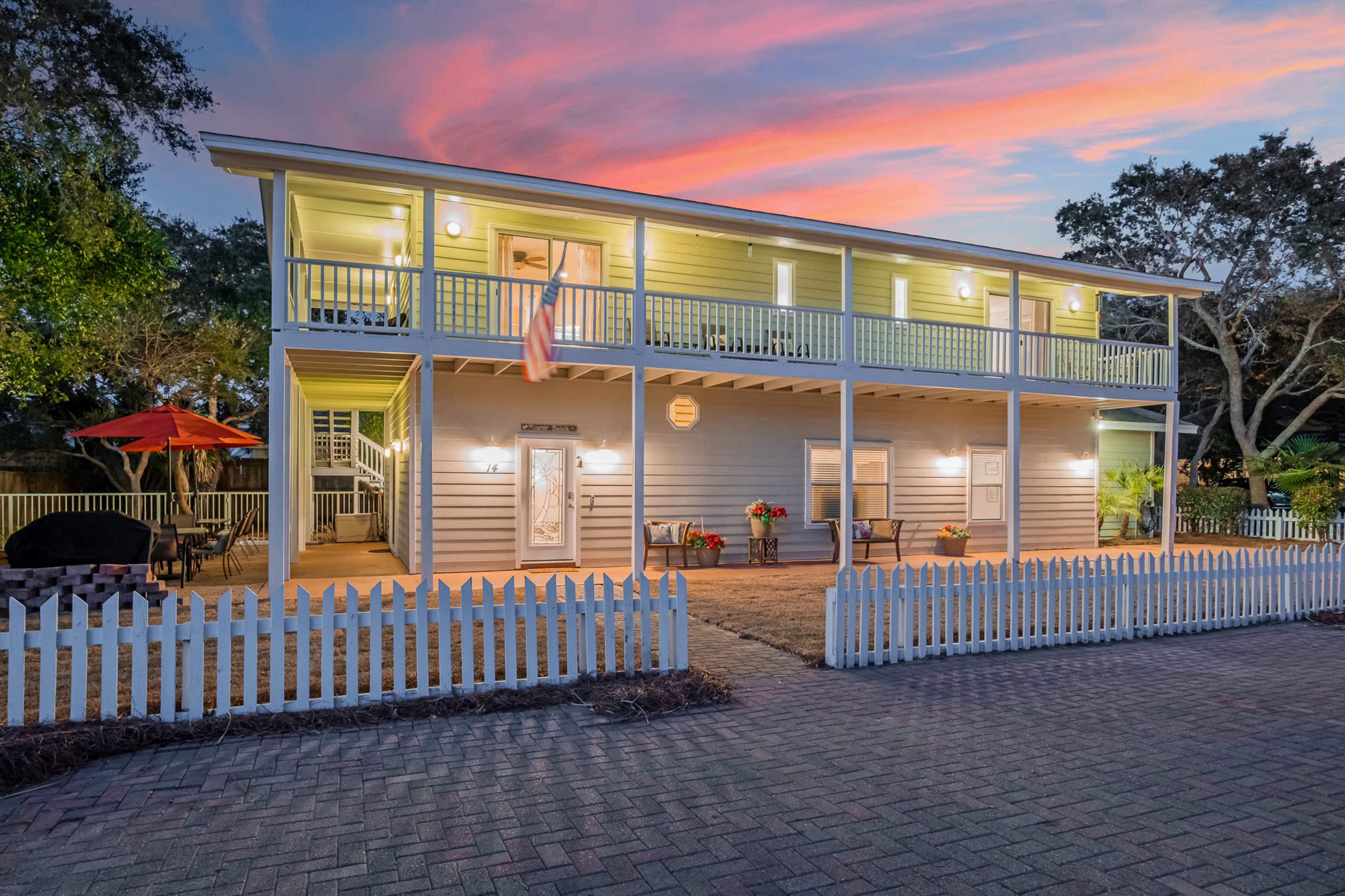 A two-story house with a front porch and a white picket fence is illuminated against a colorful sunset sky.