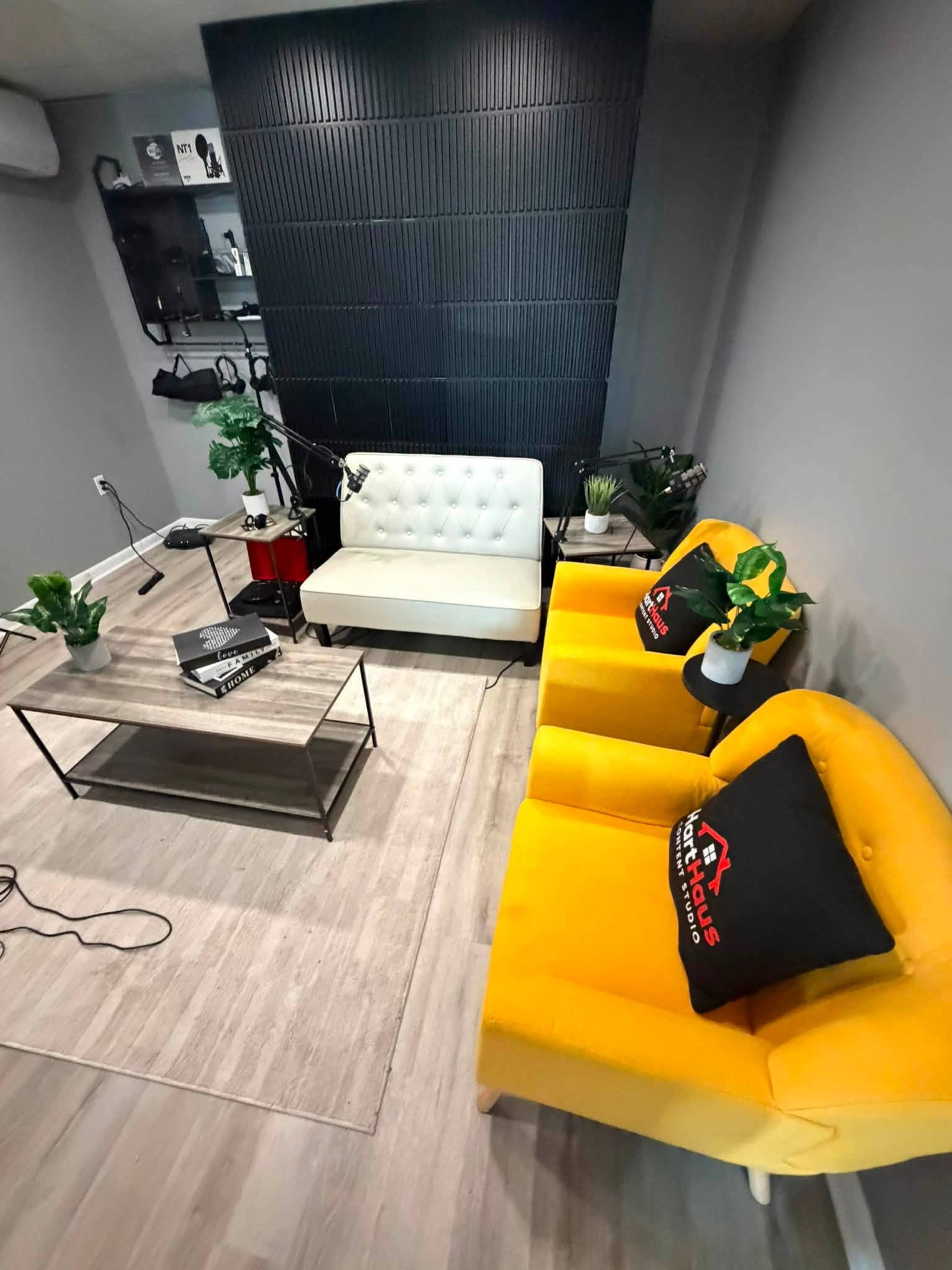 The image shows a modern interior space featuring a white sofa, two yellow armchairs, a glass coffee table, and various decorative plants against a textured black wall.