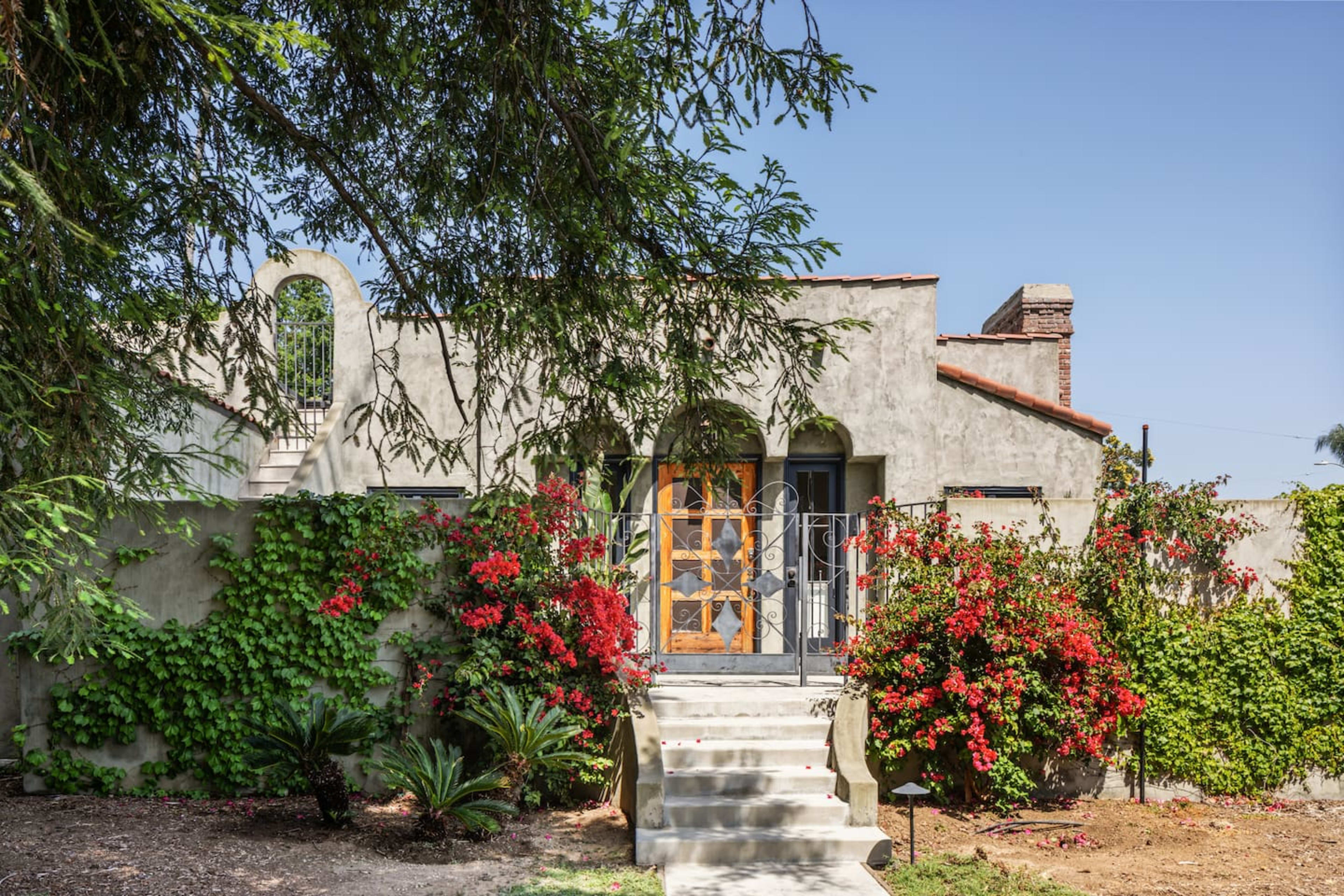 A stucco house with wooden doors is surrounded by vibrant flowering plants and has stairs leading to the entrance.