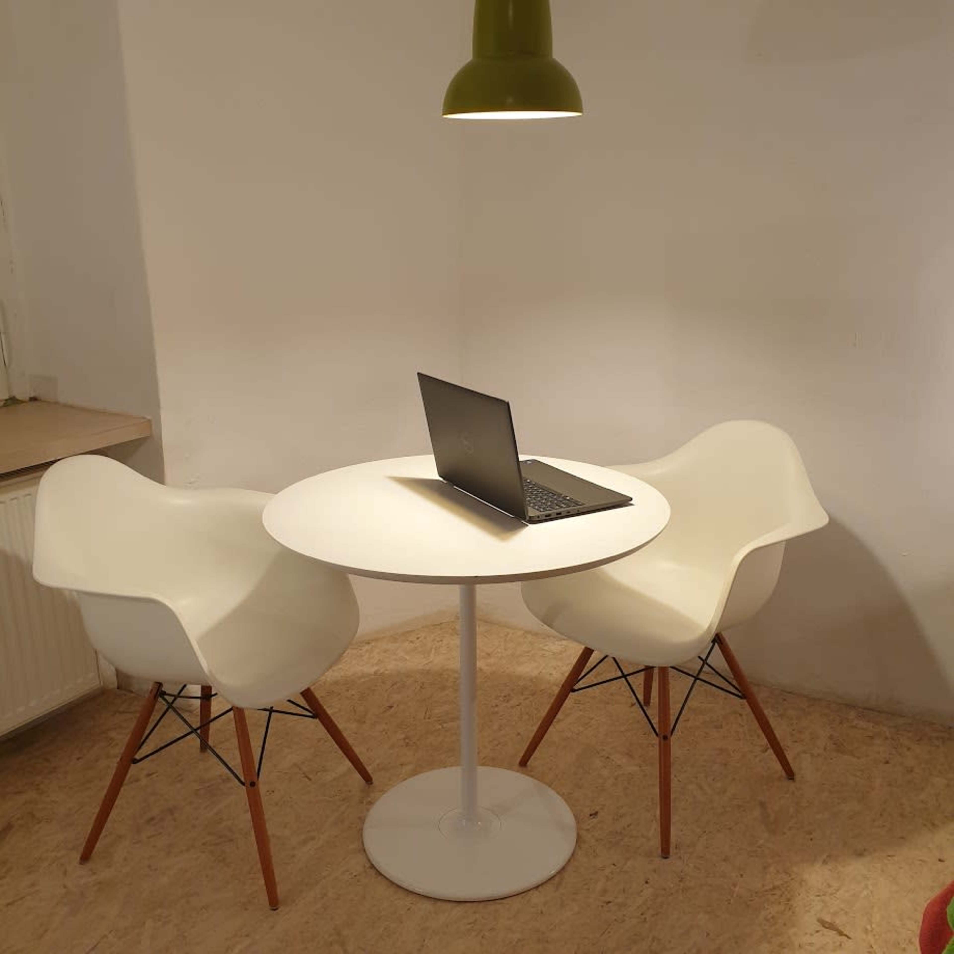 A small, round white table with a laptop sits between two modern white chairs under a green pendant light.