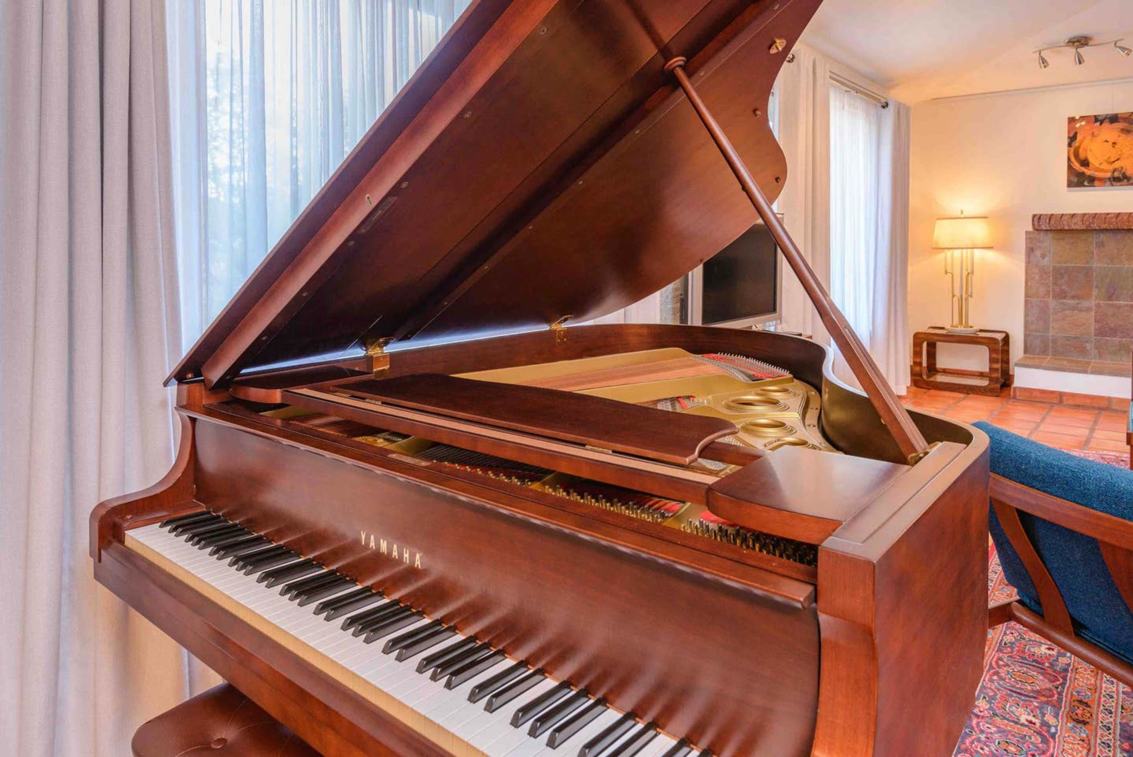 A grand piano with its lid open sits in a well-lit room, near a cozy seating area and large windows with curtains.