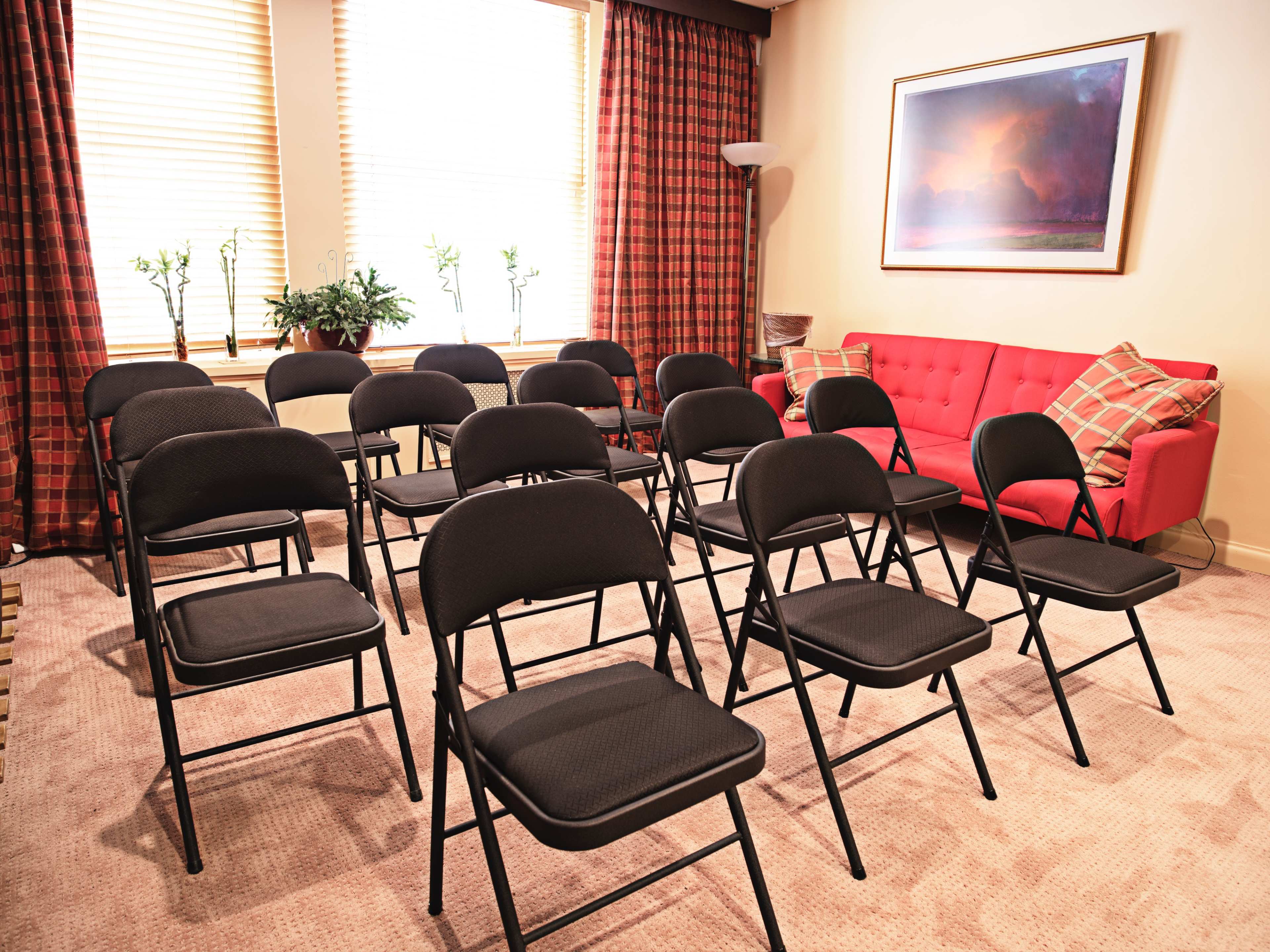 A small room features a set of black folding chairs arranged in rows facing a red couch and large windows with light filtering through the curtains.