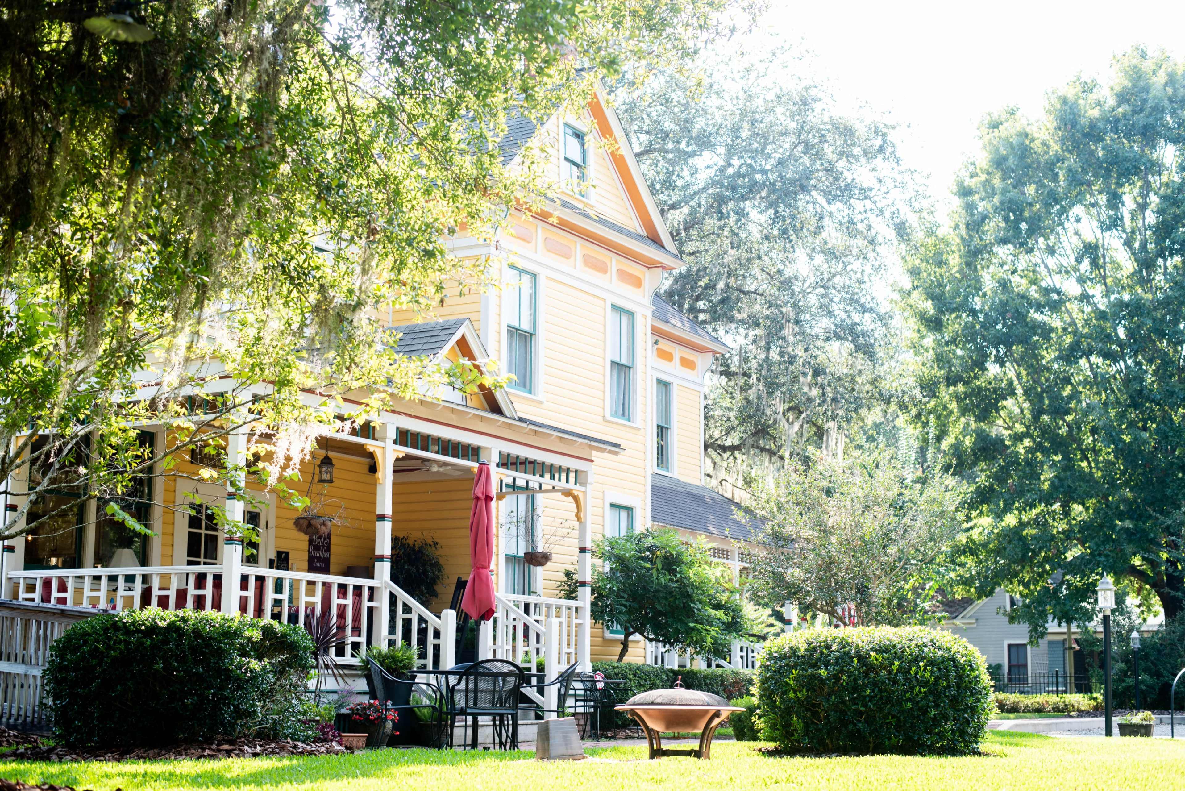 A yellow Victorian-style house stands under bright sunlight, surrounded by green trees and manicured bushes.