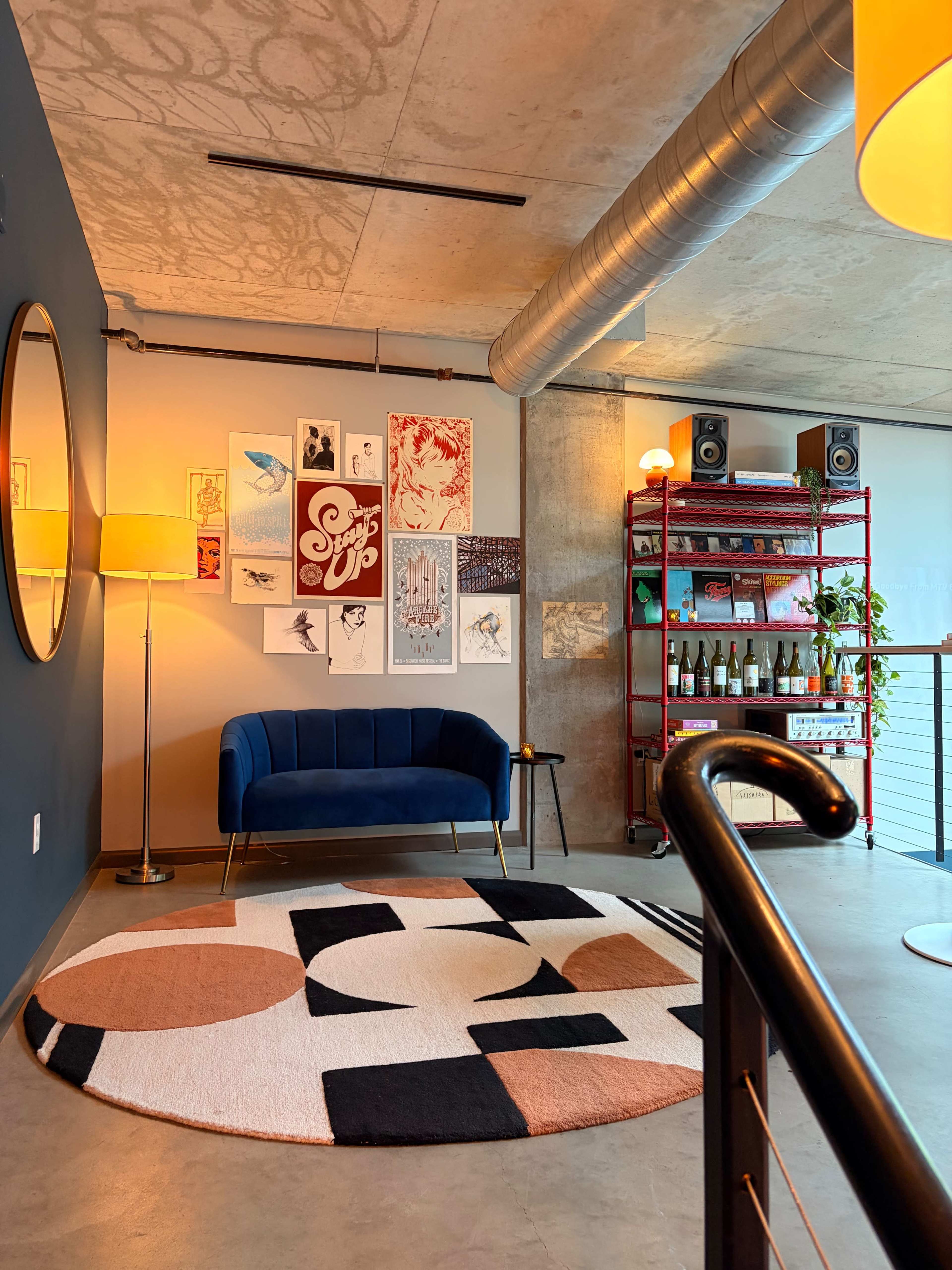 The image shows a modern interior space featuring a blue velvet sofa, a round rug with geometric patterns, and a collection of framed artworks on the wall.