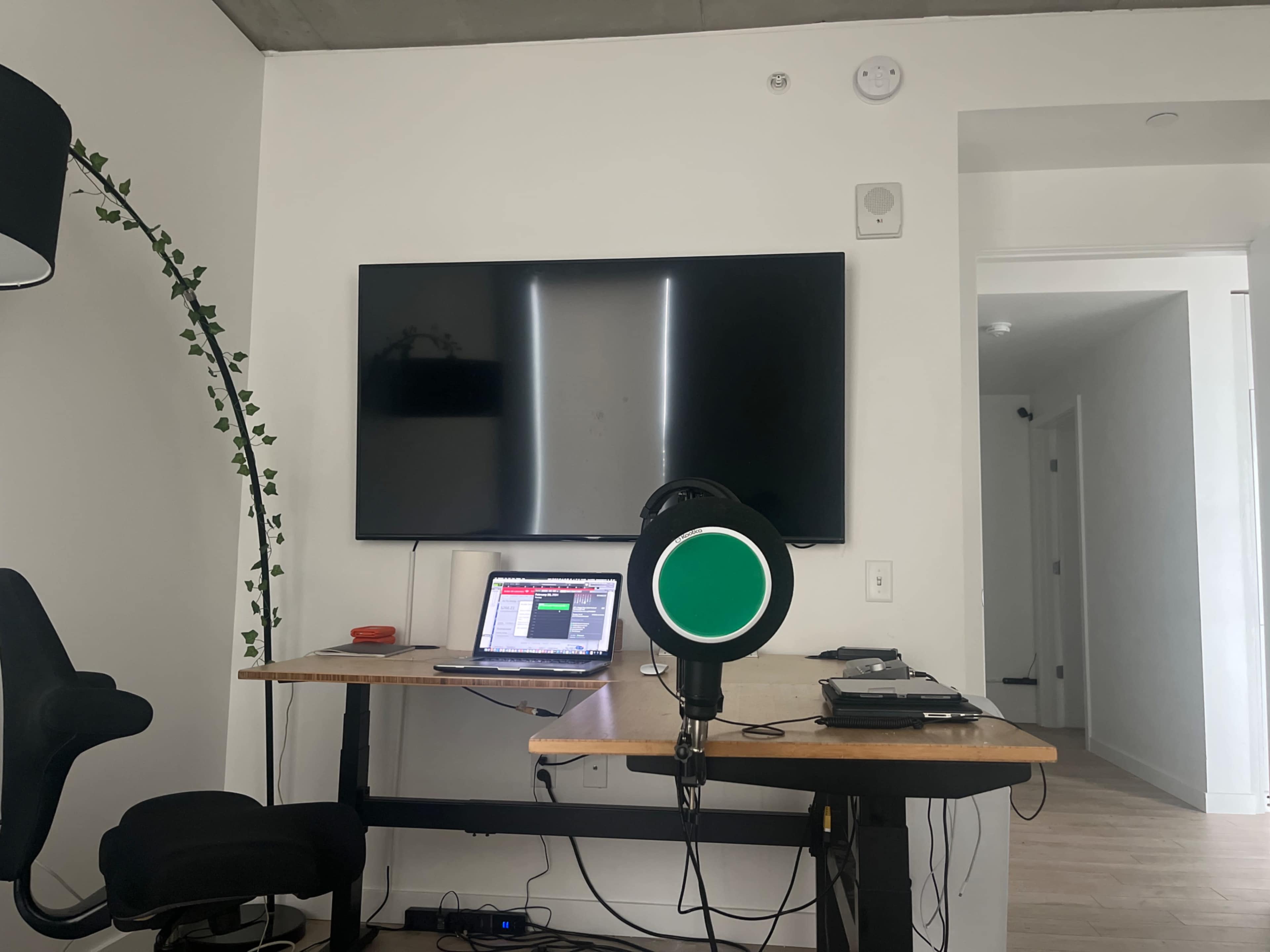 A desk with a laptop and microphone is positioned in front of a large flat-screen TV in a minimalist room.