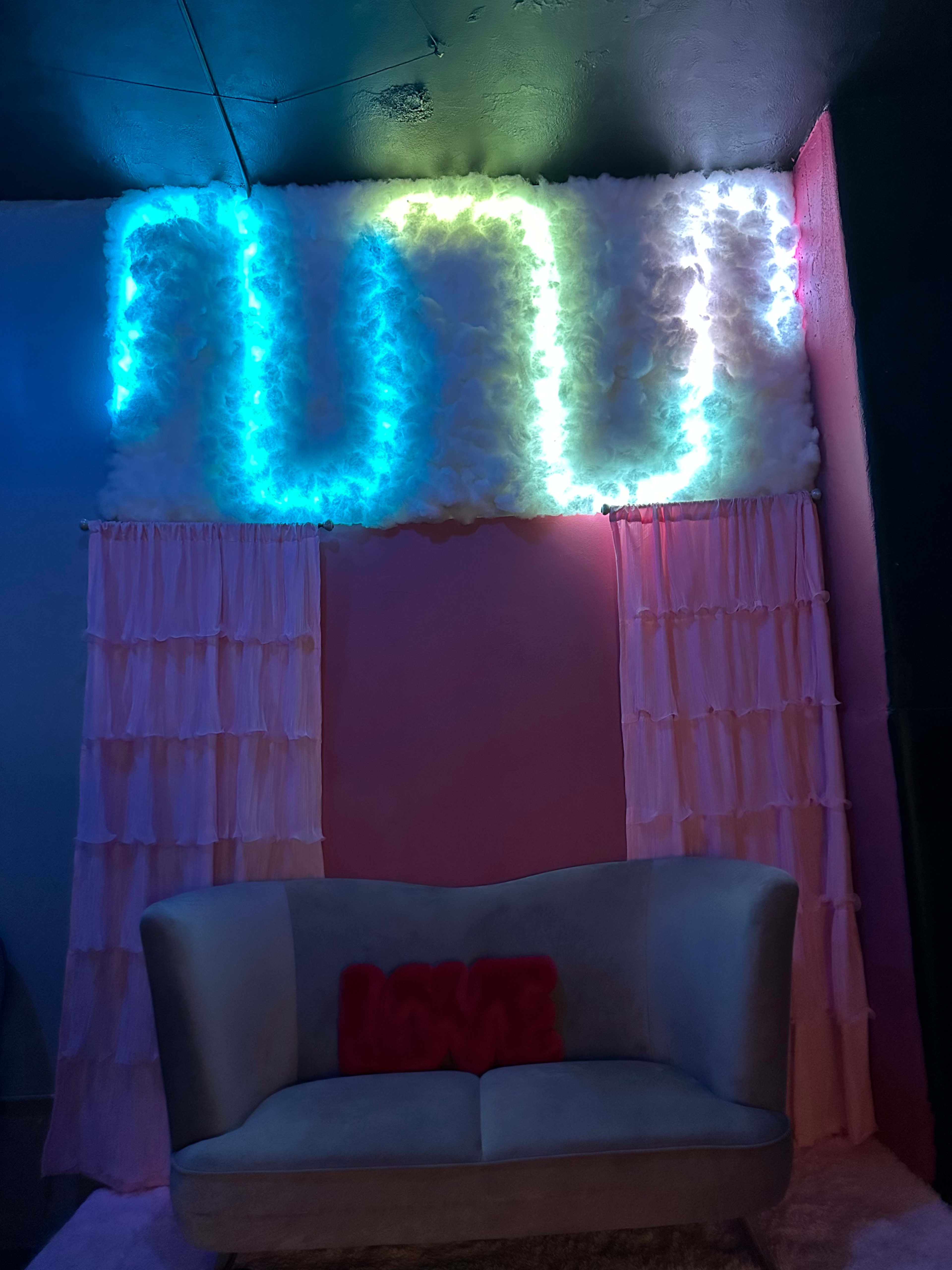 A cozy lounge area with a gray couch and illuminated cloud-like decorations above it, surrounded by soft, pink curtains.