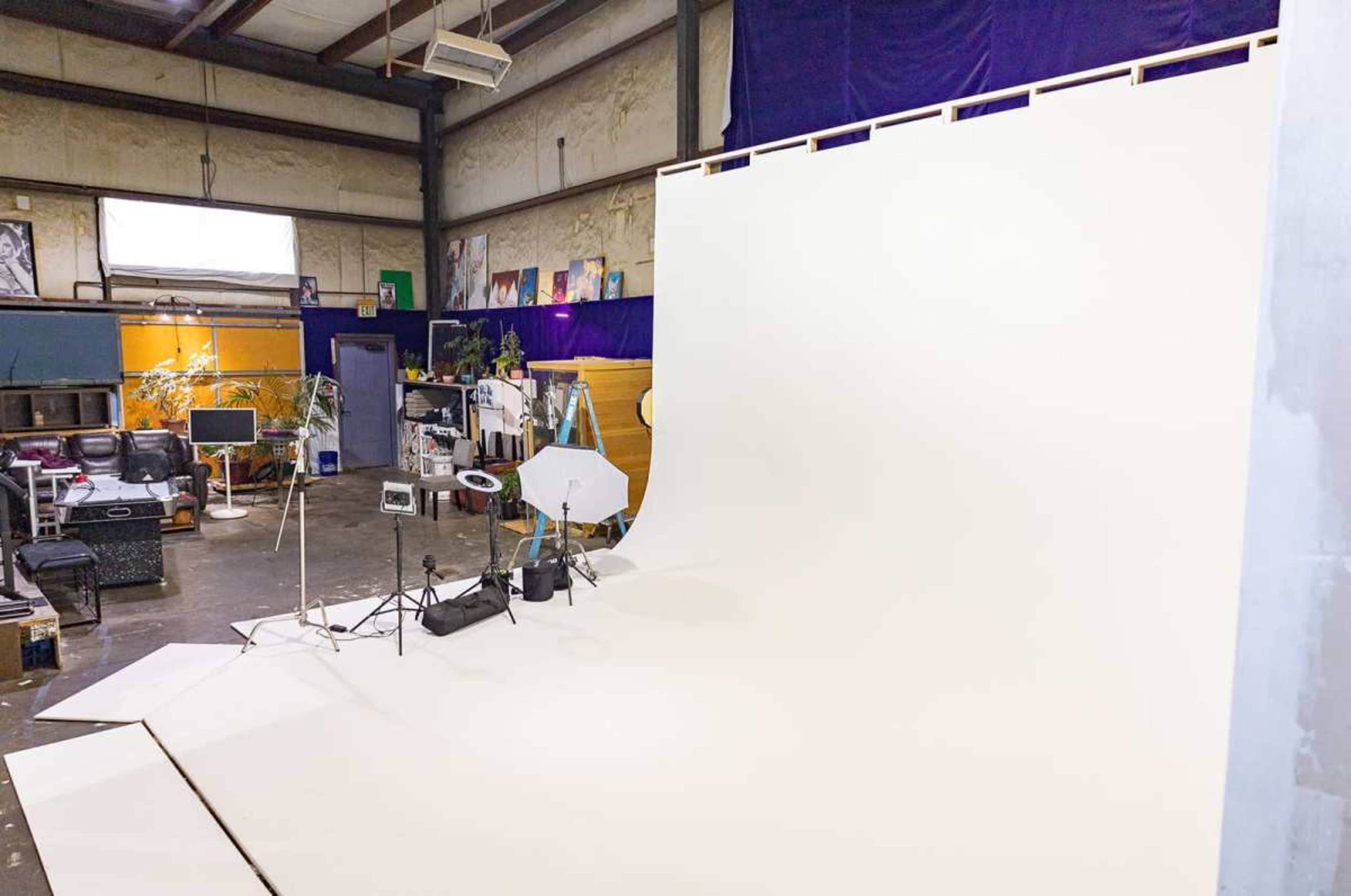 The image shows a spacious photography studio with a large white backdrop, lighting equipment, and various props organized around the room.
