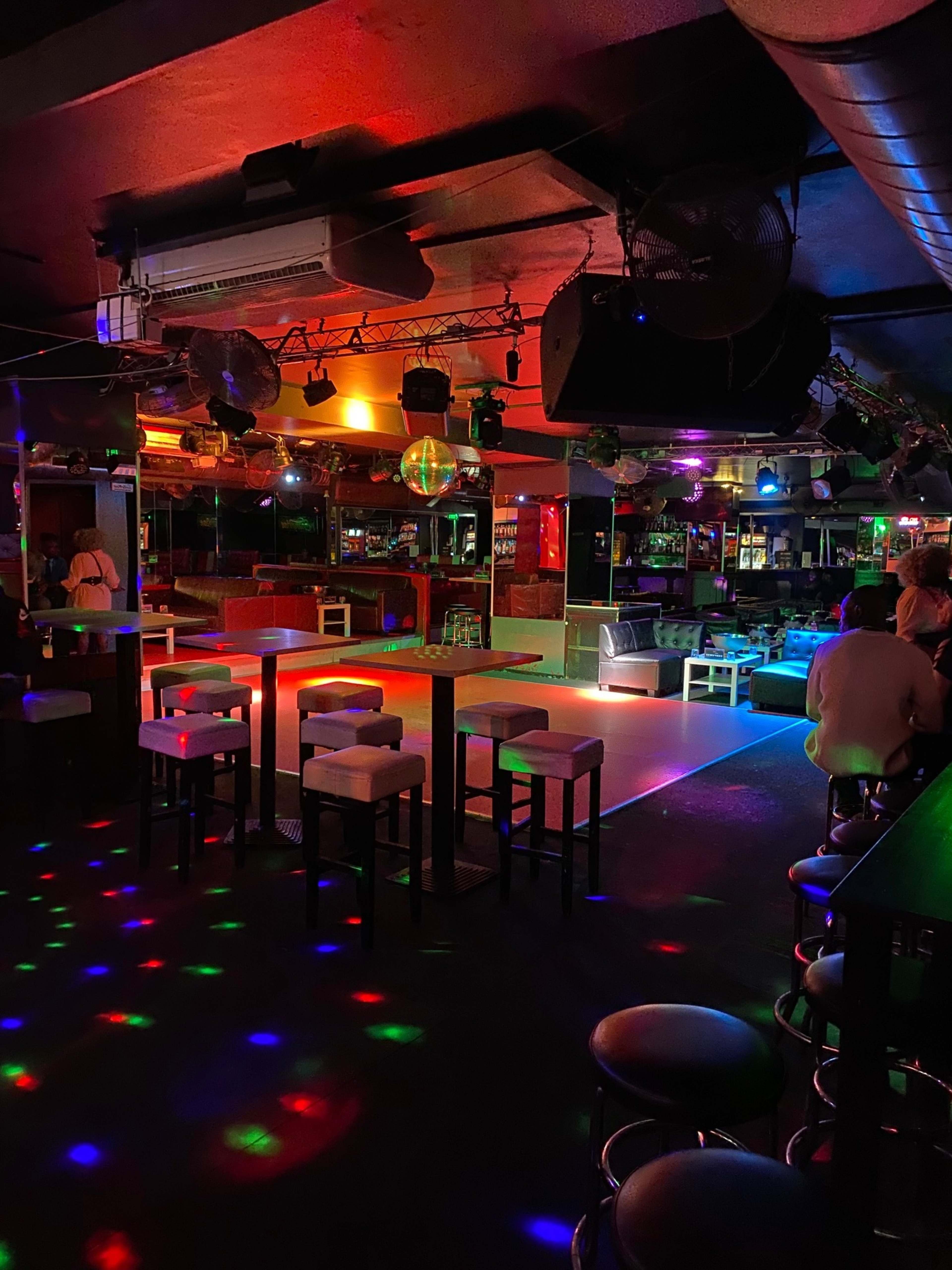 A dimly lit bar features colorful lights and a dance floor surrounded by stools and tables.