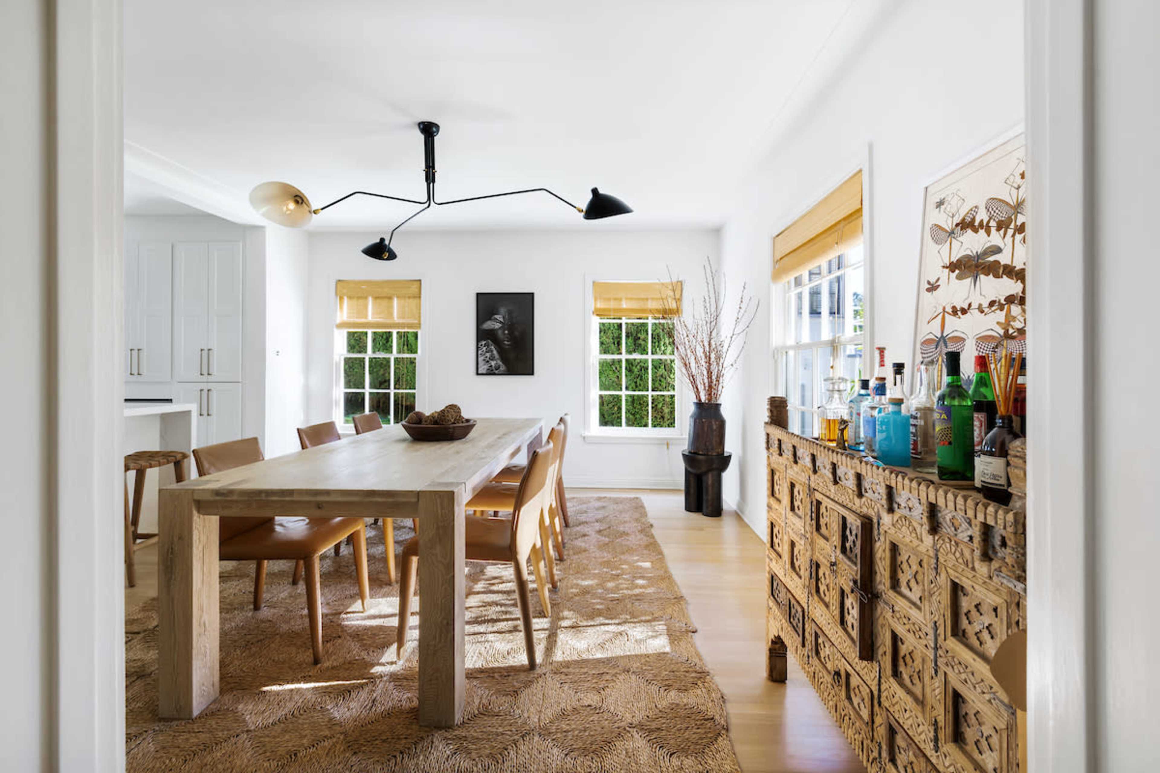 A bright dining area features a wooden table surrounded by chairs, complemented by a decorative cabinet and large windows with shades.