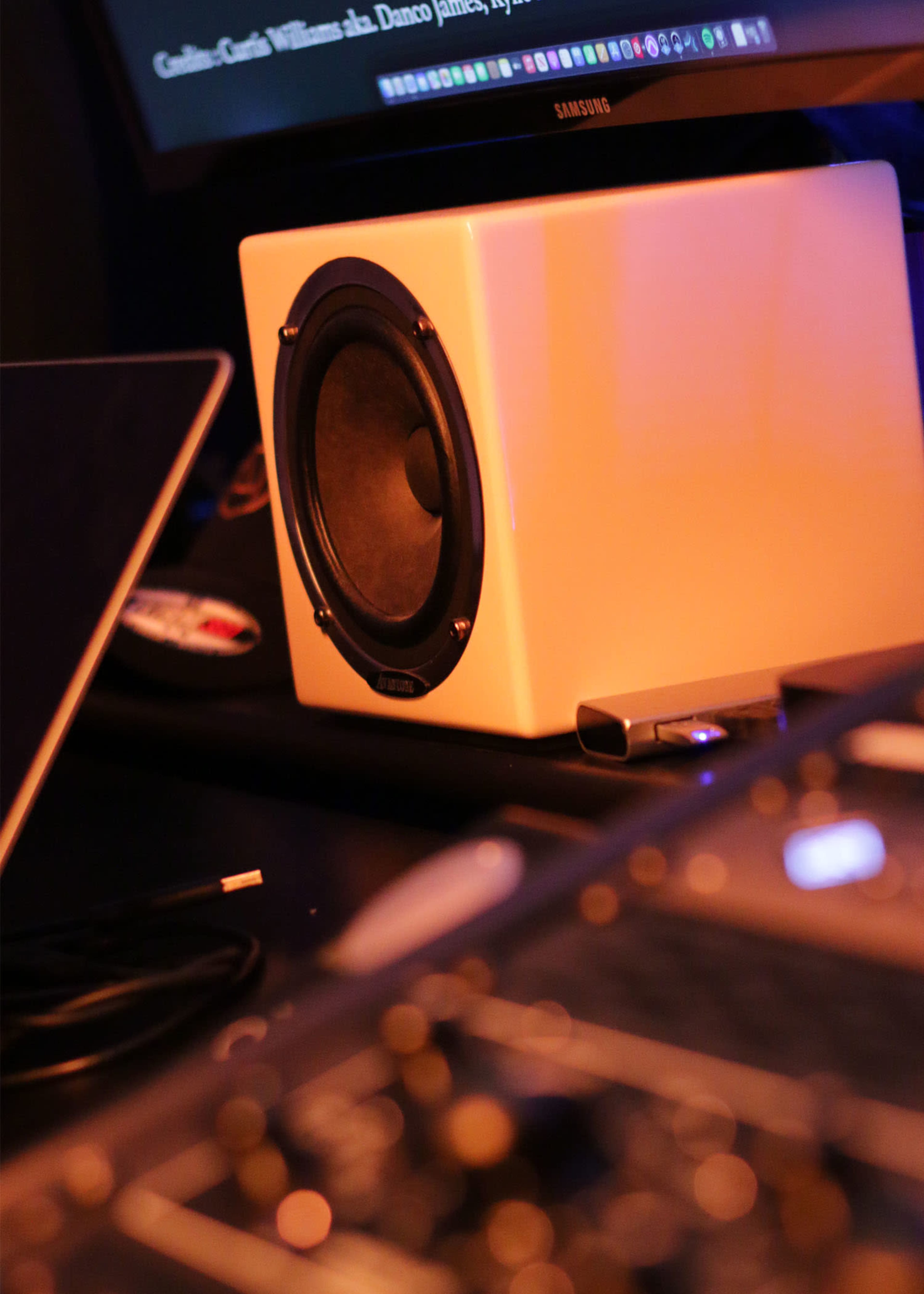 A white speaker sits on a desk next to a laptop, with a computer monitor displaying text in the background.